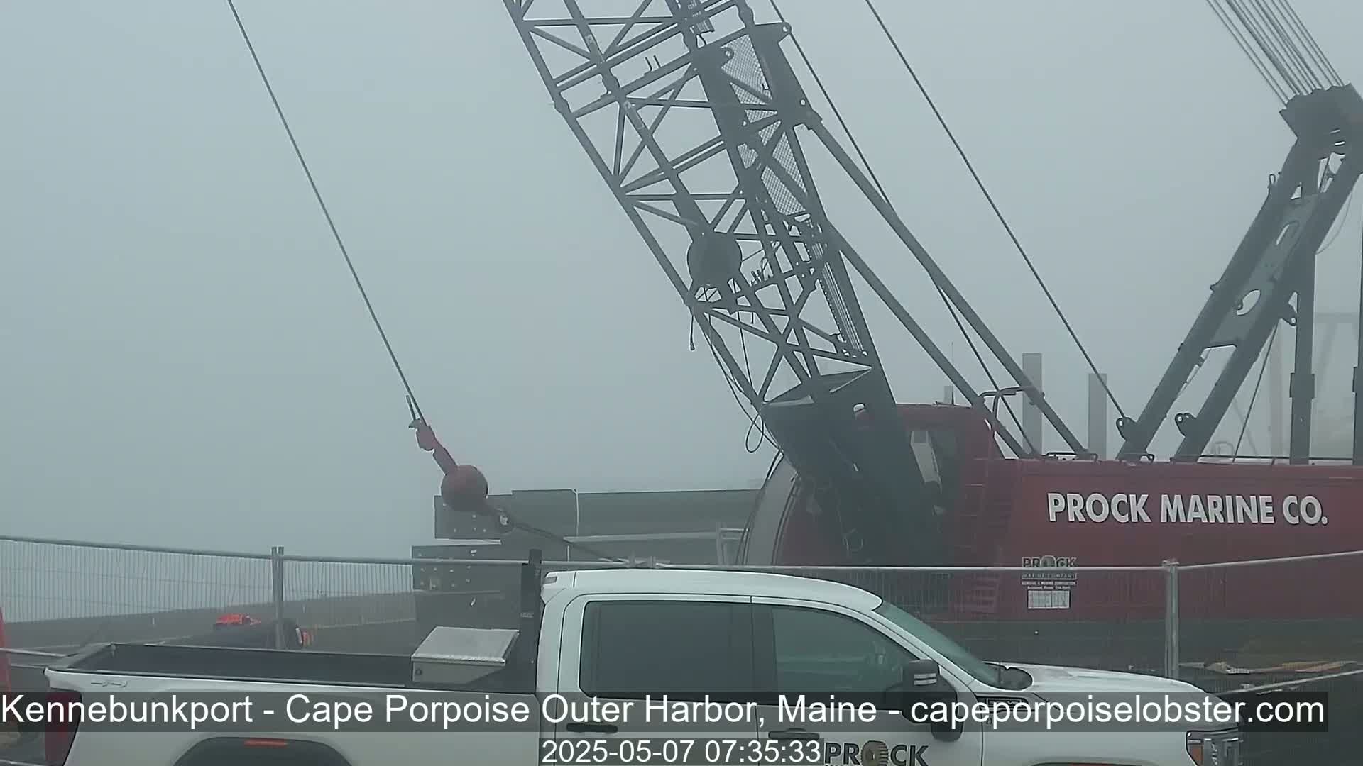 A large red crane with a cable extending towards a white pickup truck is visible in foggy conditions.