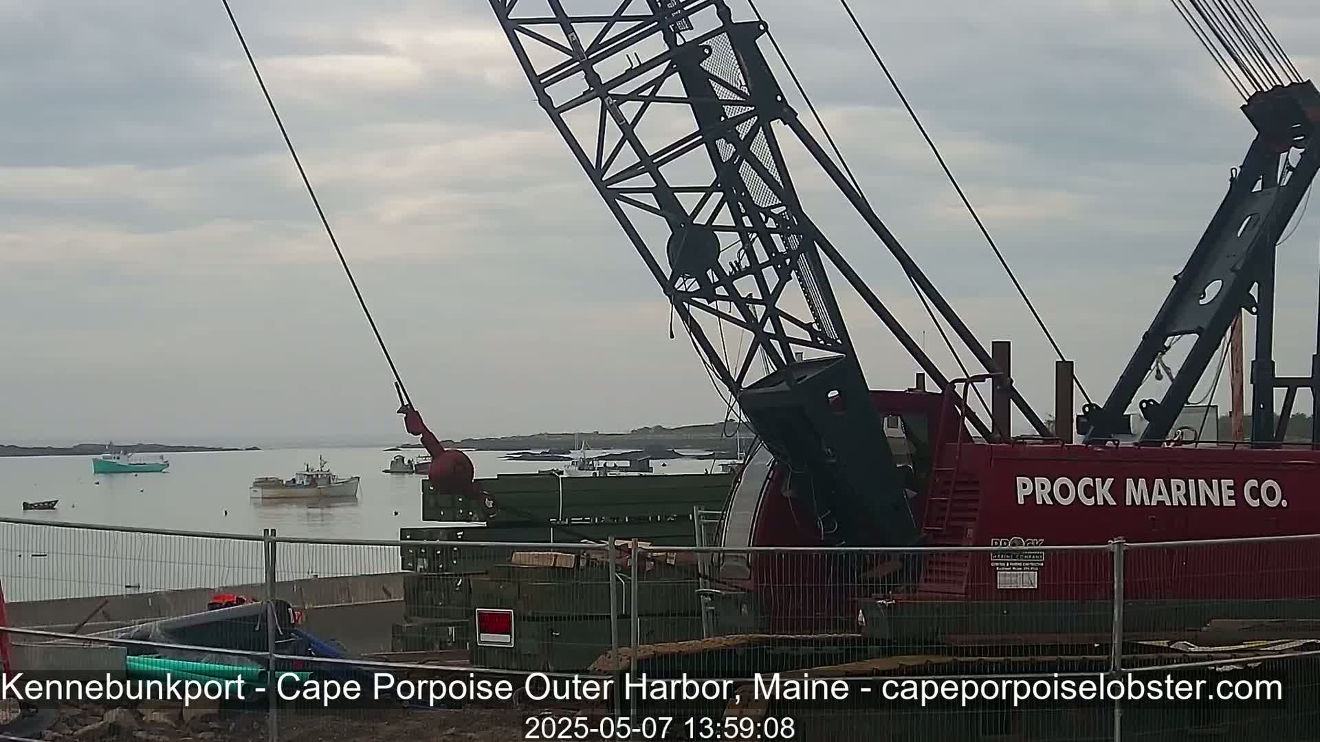 A large red crane is positioned near a harbor with several small boats, under an overcast sky.