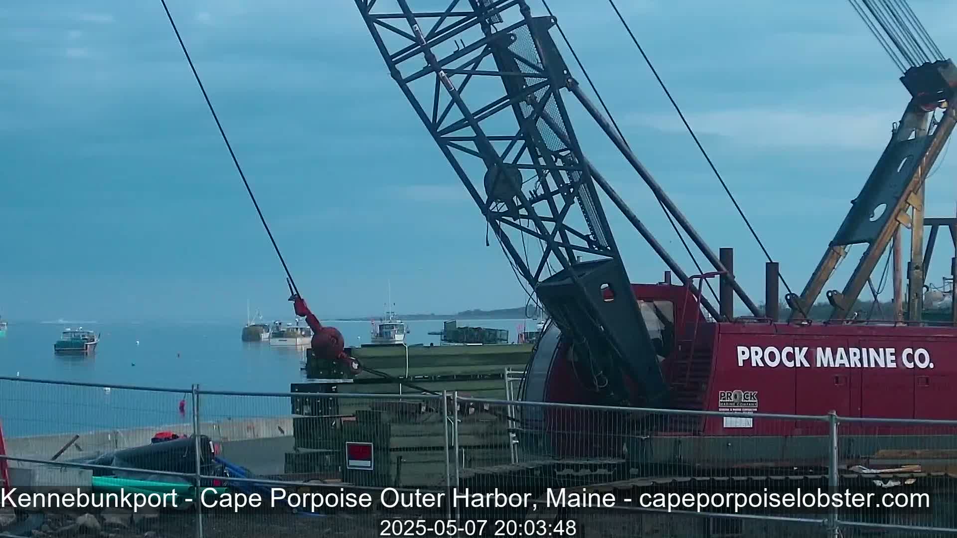 A red crane is positioned near a harbor on a clear day, with several boats visible in the calm water.
