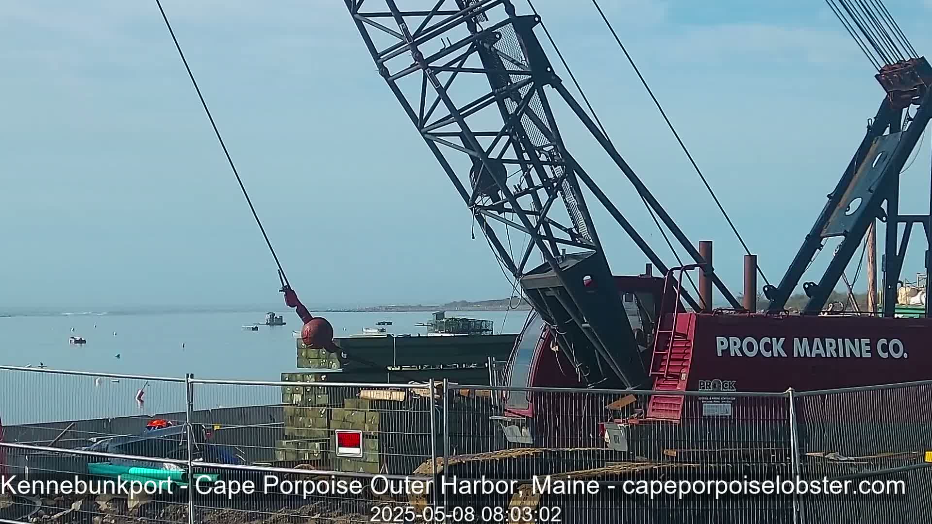 A large red crane is positioned near a body of water on a partly sunny day, with several small boats visible in the distance.