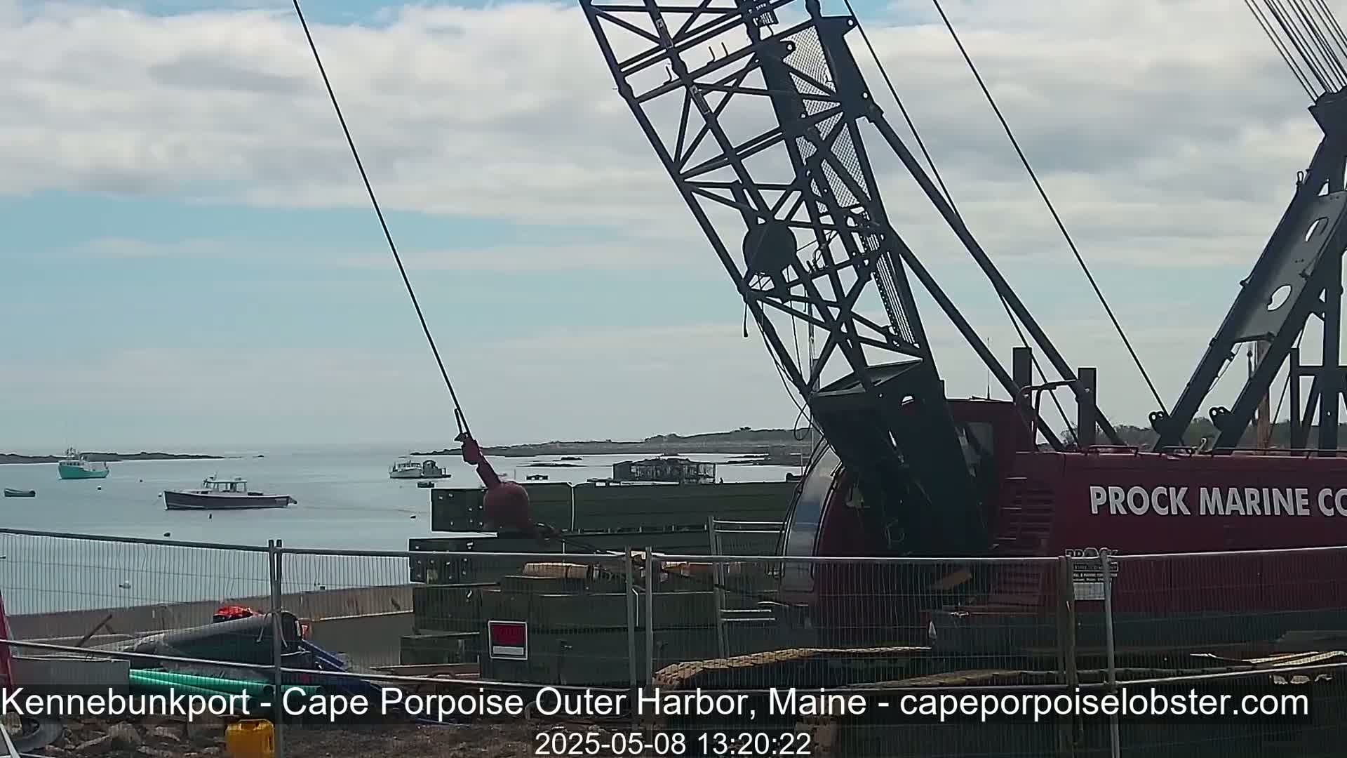 A large red crane is positioned near a body of water where several small boats are visible under a partly cloudy sky.