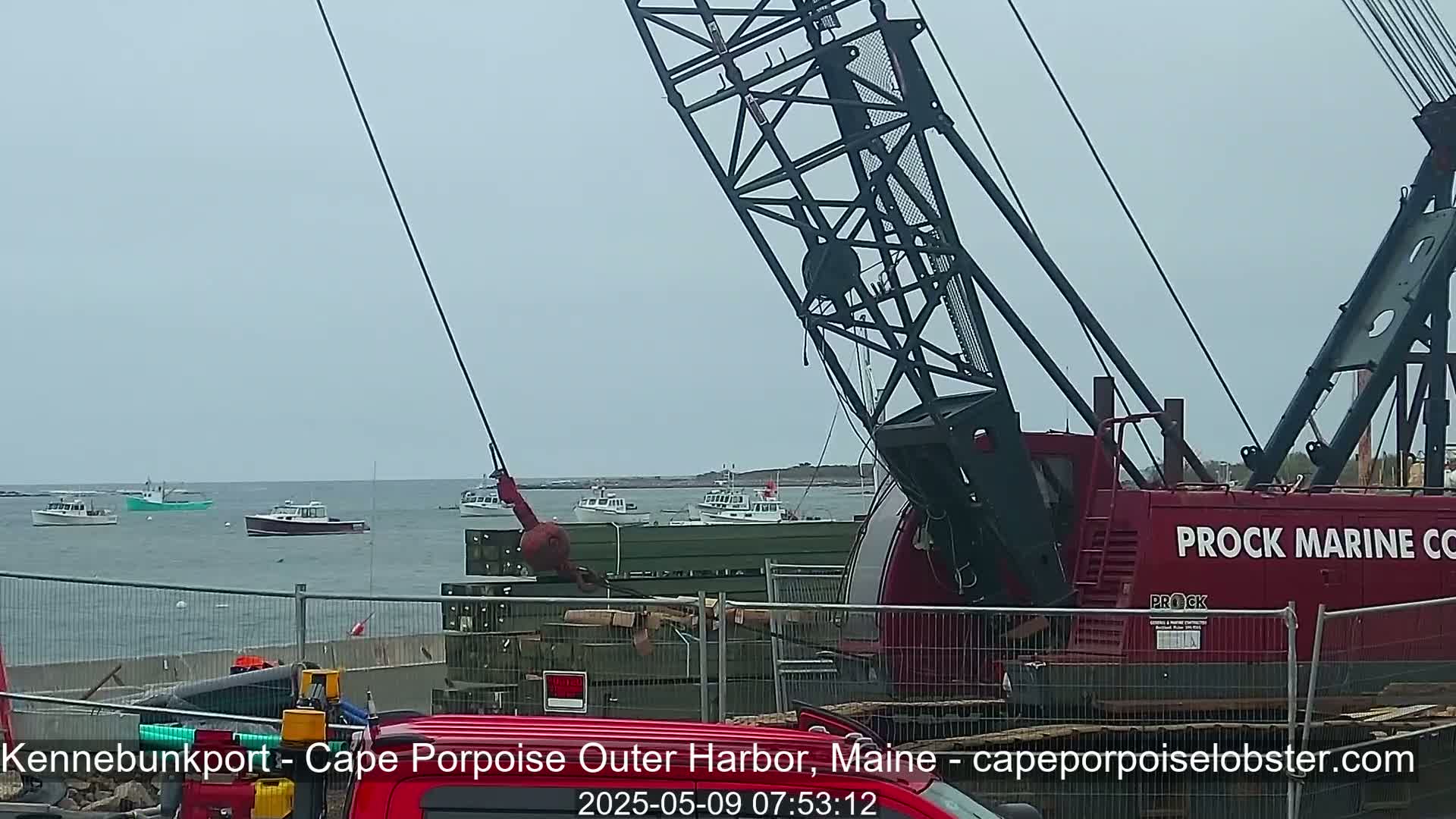 A red crane is positioned near a harbor on an overcast day, with several boats visible in the background.