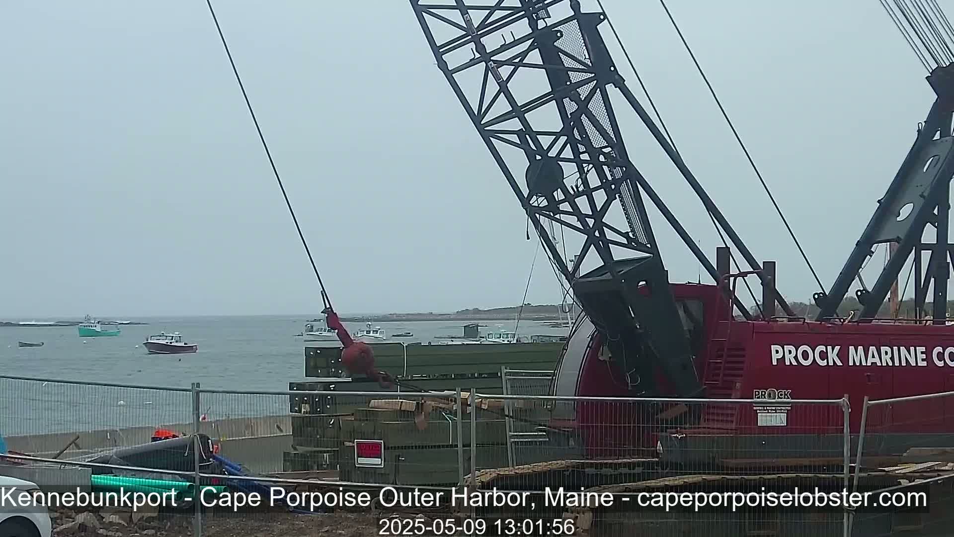 A large red crane is lifting a heavy object near a harbor on an overcast day, with several small boats visible in the distance.
