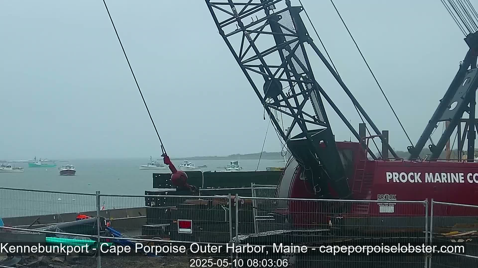 A large red crane is positioned near a harbor on an overcast day, with several small fishing boats visible in the distance.