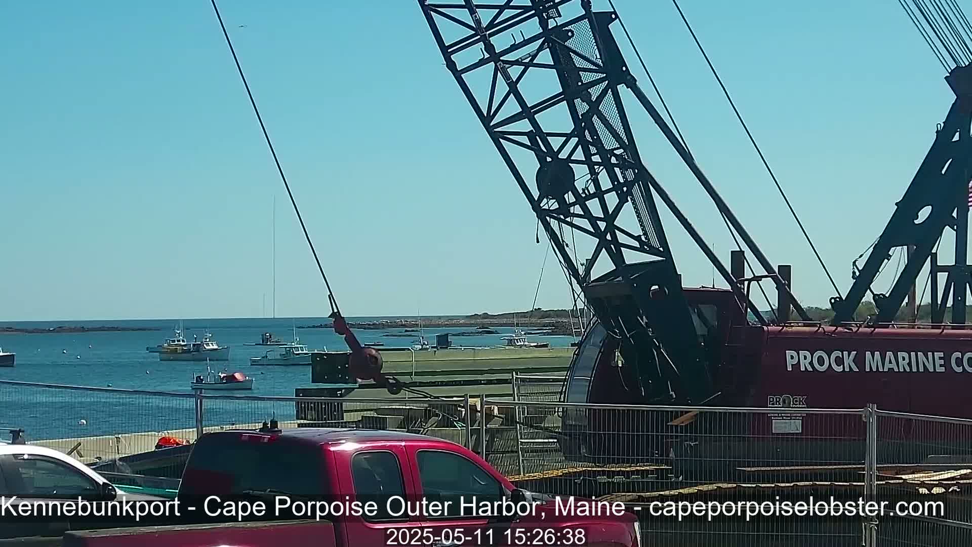 A large red crane is positioned near a harbor on a sunny day, with several small fishing boats visible in the calm water.