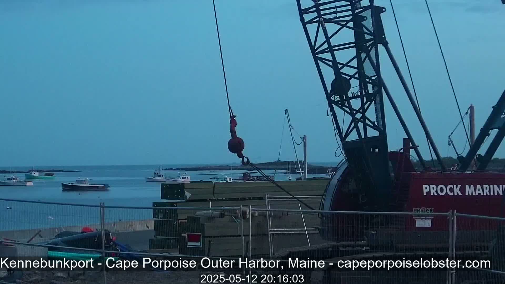A large red crane with a hook hangs over a harbor at dusk, where several small boats are moored near a construction site.