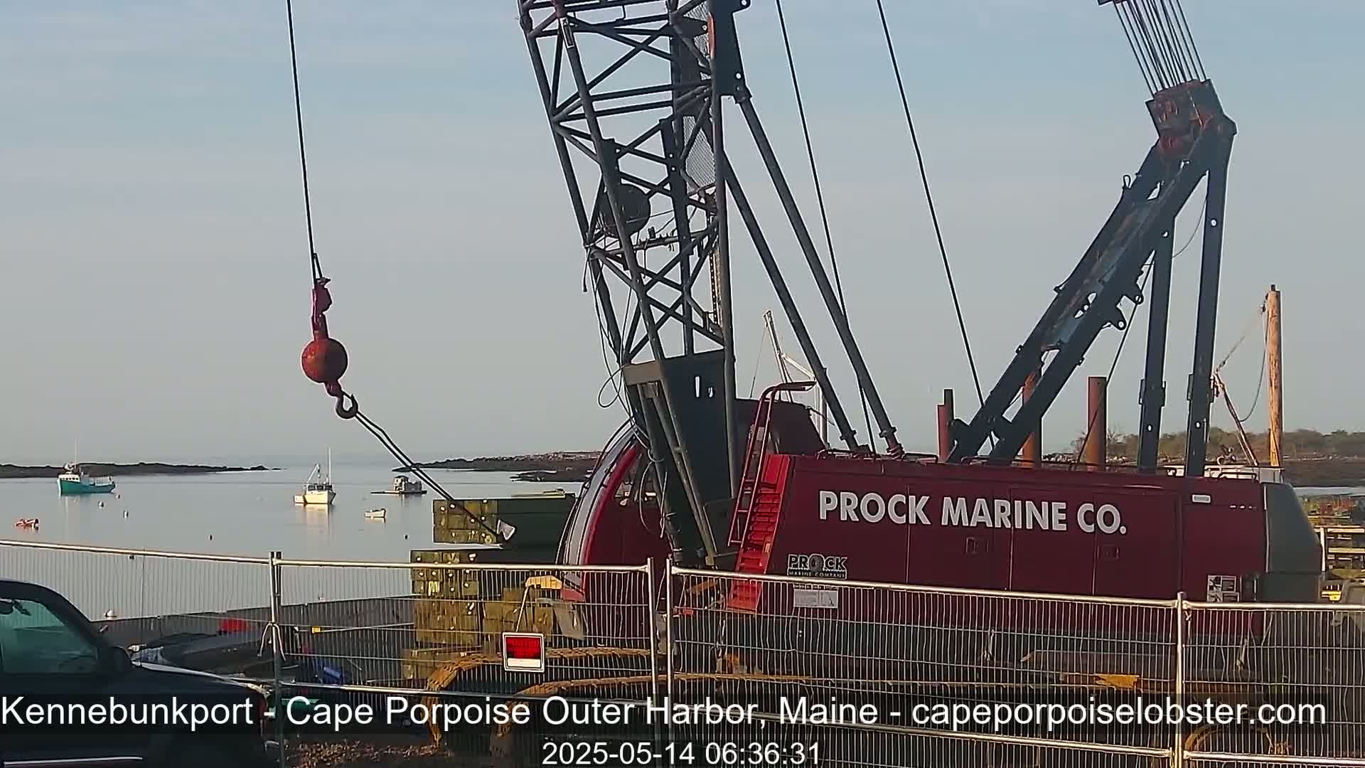 A red Prock Marine Co. crane with a large hook is positioned near a calm body of water where several small boats are moored, under a clear sky.