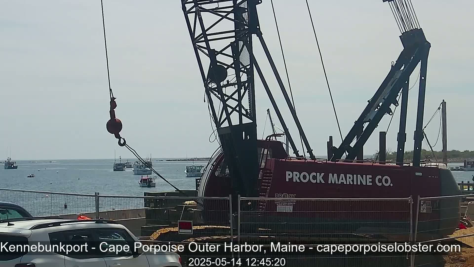 A large red crane with a hook is positioned near a body of water where several small boats are visible on a partly sunny day.