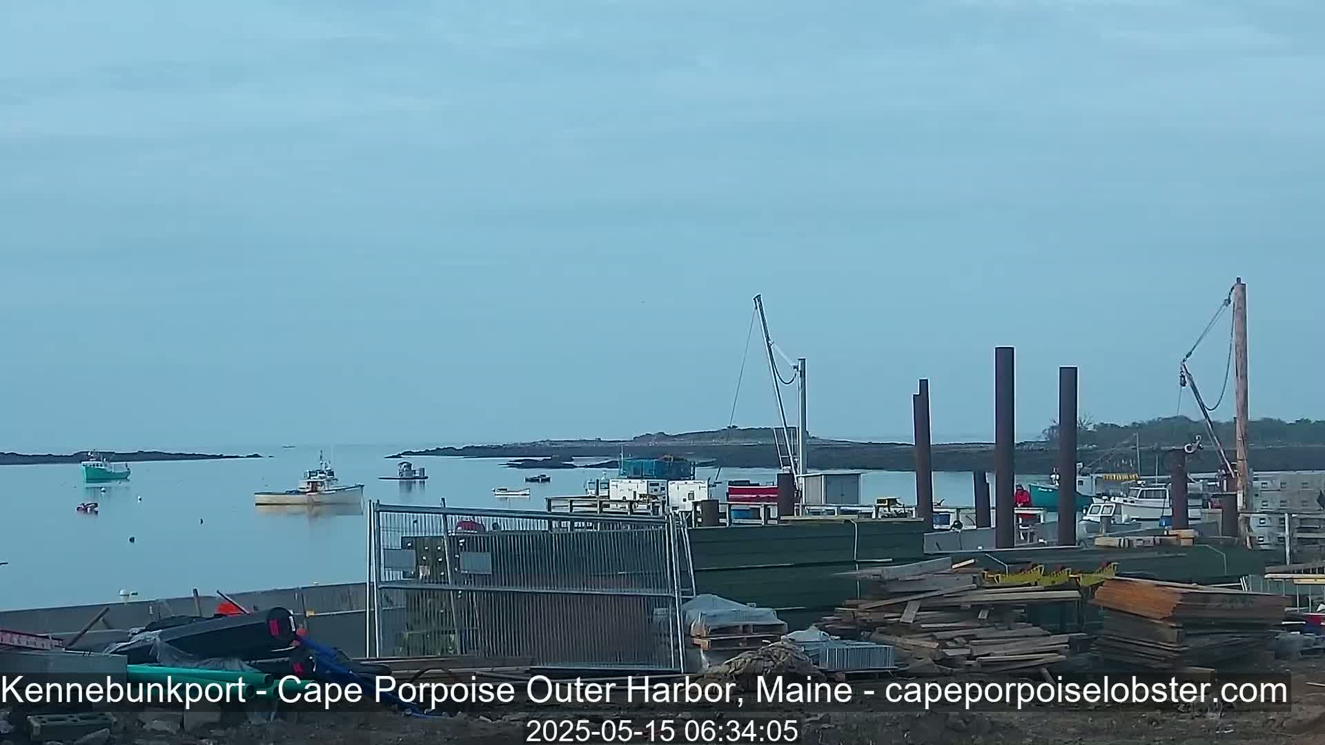 Several boats are moored in a calm harbor under a clear, light-blue sky, with construction materials and equipment visible on the shore.