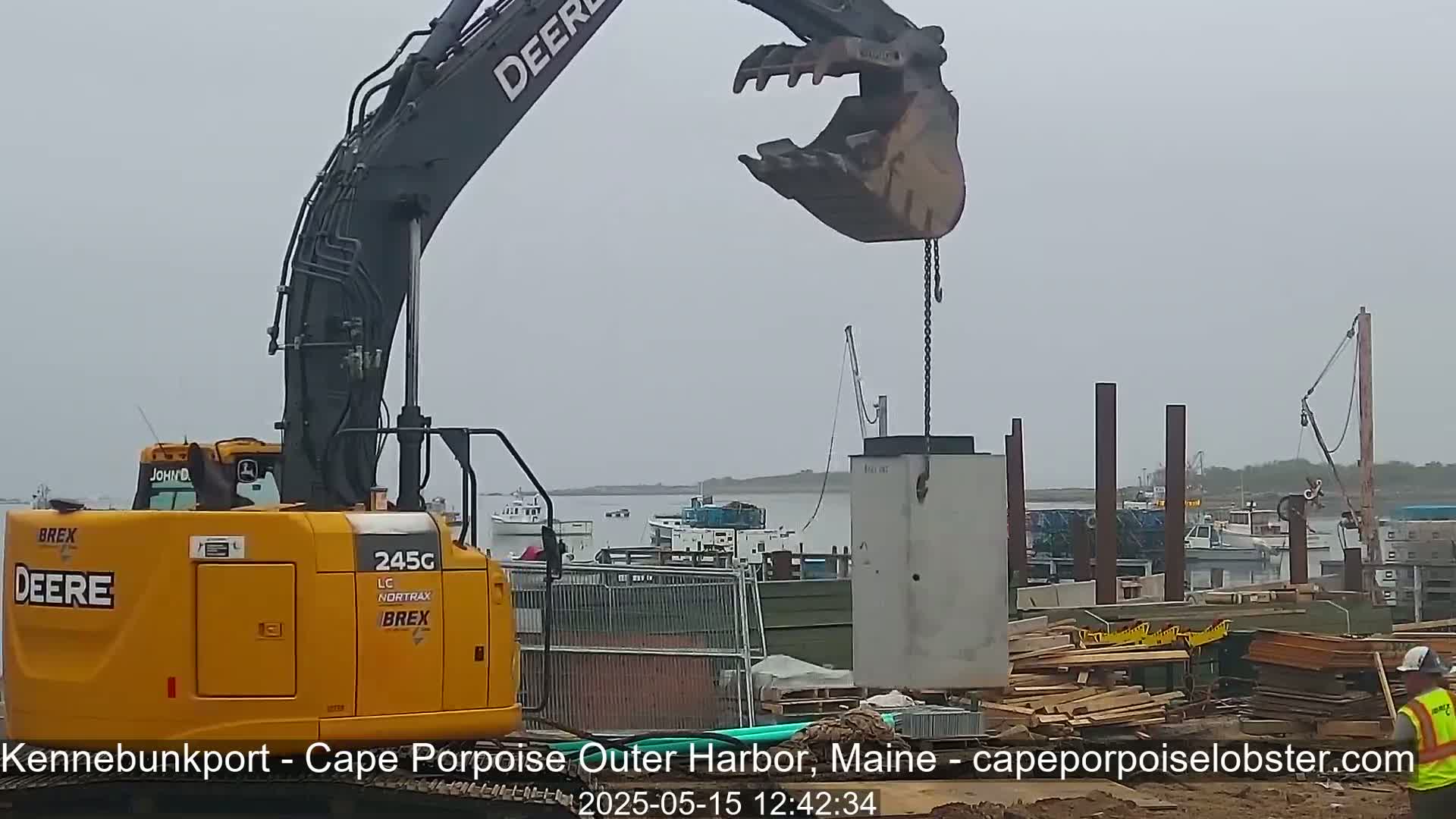 A yellow excavator lifts a large concrete block near a harbor on an overcast day.