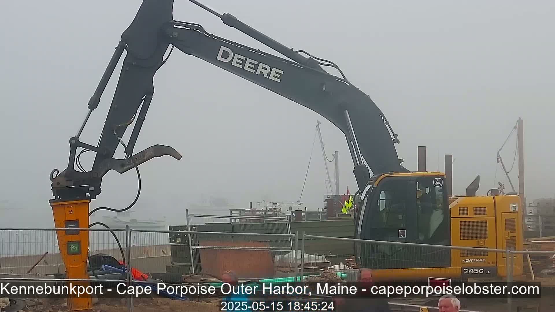 A large excavator with a demolition hammer attached is operating near a harbor in foggy conditions.