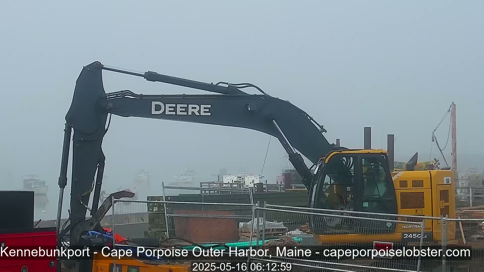 A large Deere excavator sits on a foggy construction site near a harbor with several boats visible in the distance.