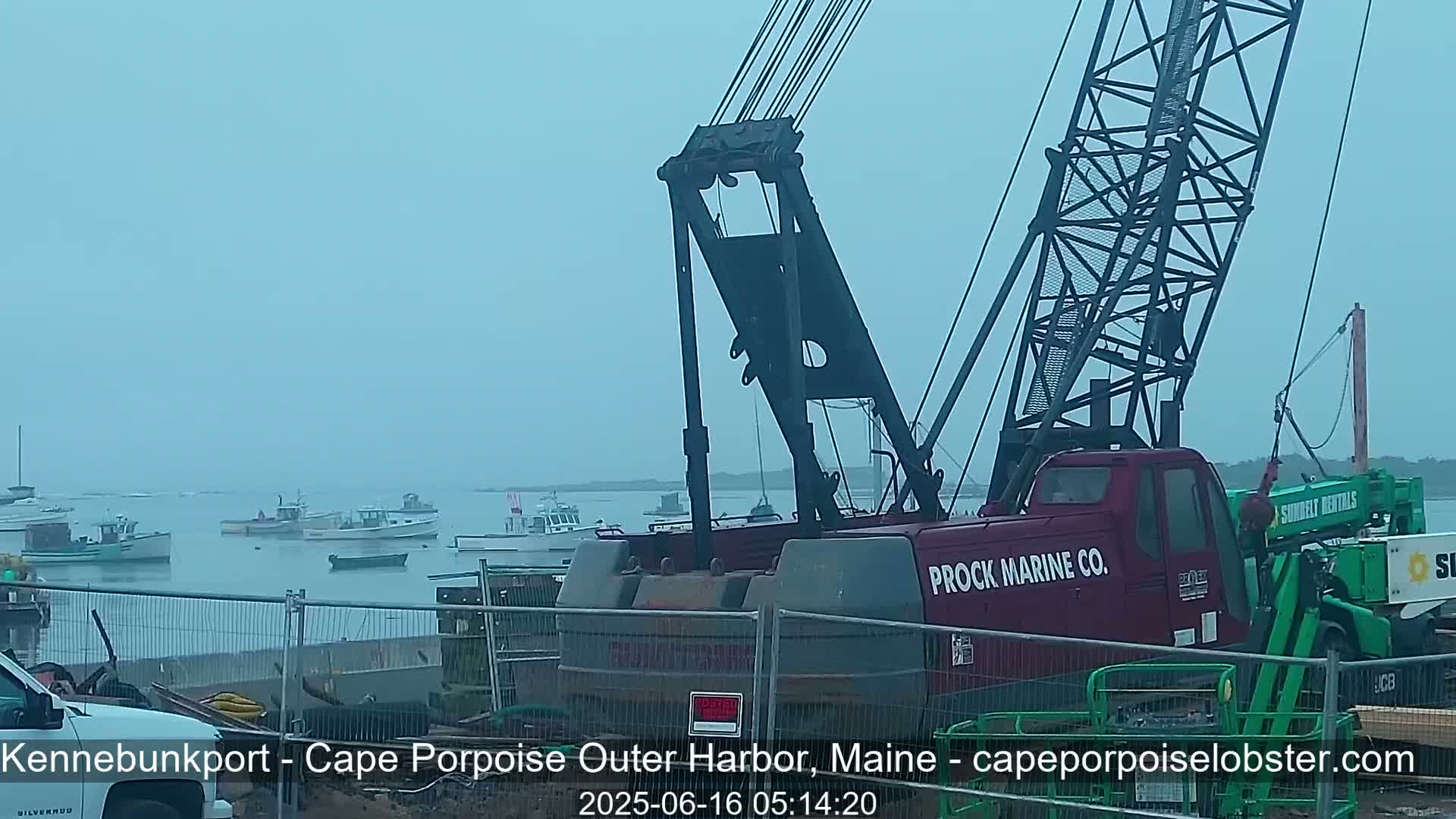 A large red crane sits in a harbor on an overcast day, with numerous smaller fishing boats visible in the background.