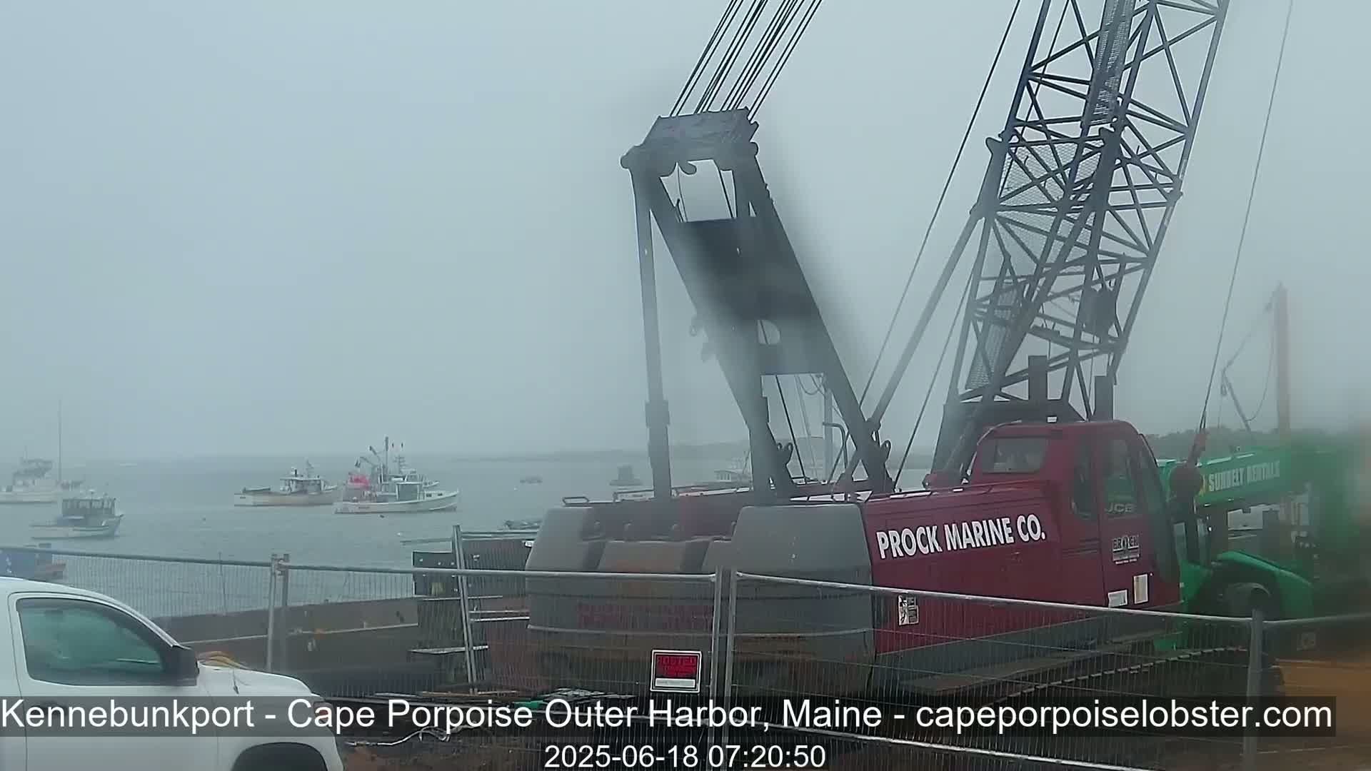 A large red crane sits in a harbor on a foggy day, with several fishing boats visible in the background.