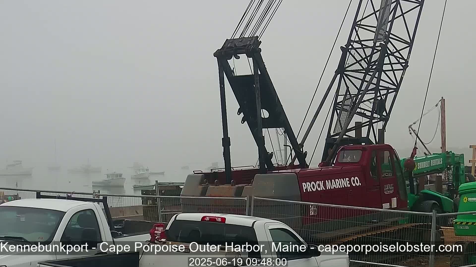 A large red crane sits near several parked trucks in a foggy harbor, with several boats barely visible in the distance.