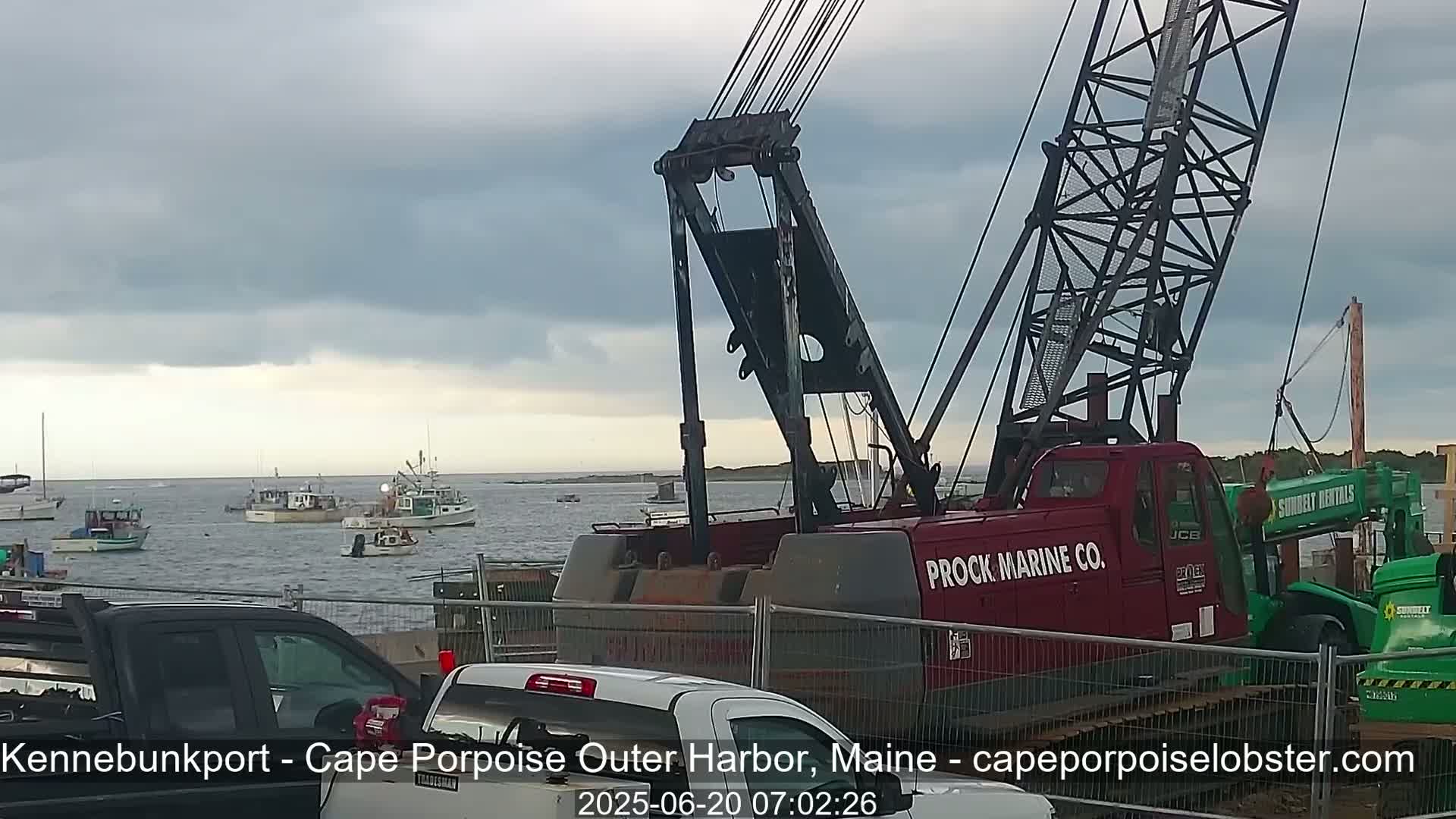 A red Prock Marine Co. crane sits near a harbor with several boats under a cloudy sky.