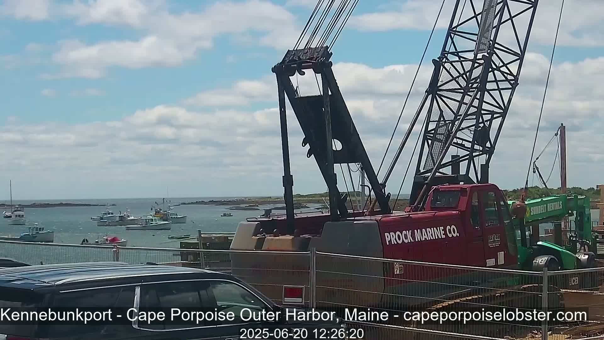 A large red crane is positioned near a harbor filled with several small boats under a partly cloudy sky.