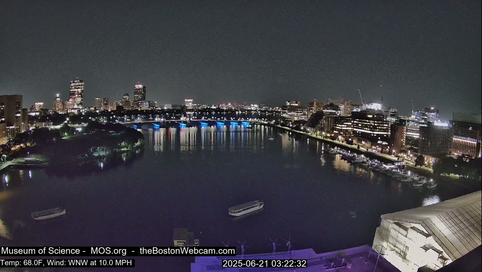 A nighttime view of a city skyline across a dark river, with a bridge illuminated in blue, under a clear sky.