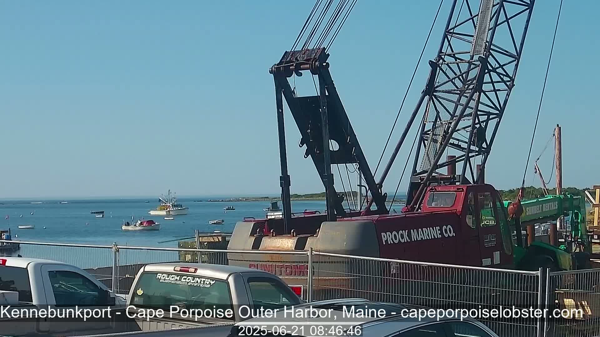 A large red crane is parked near a harbor on a sunny day, with several fishing boats visible on the calm water in the background.