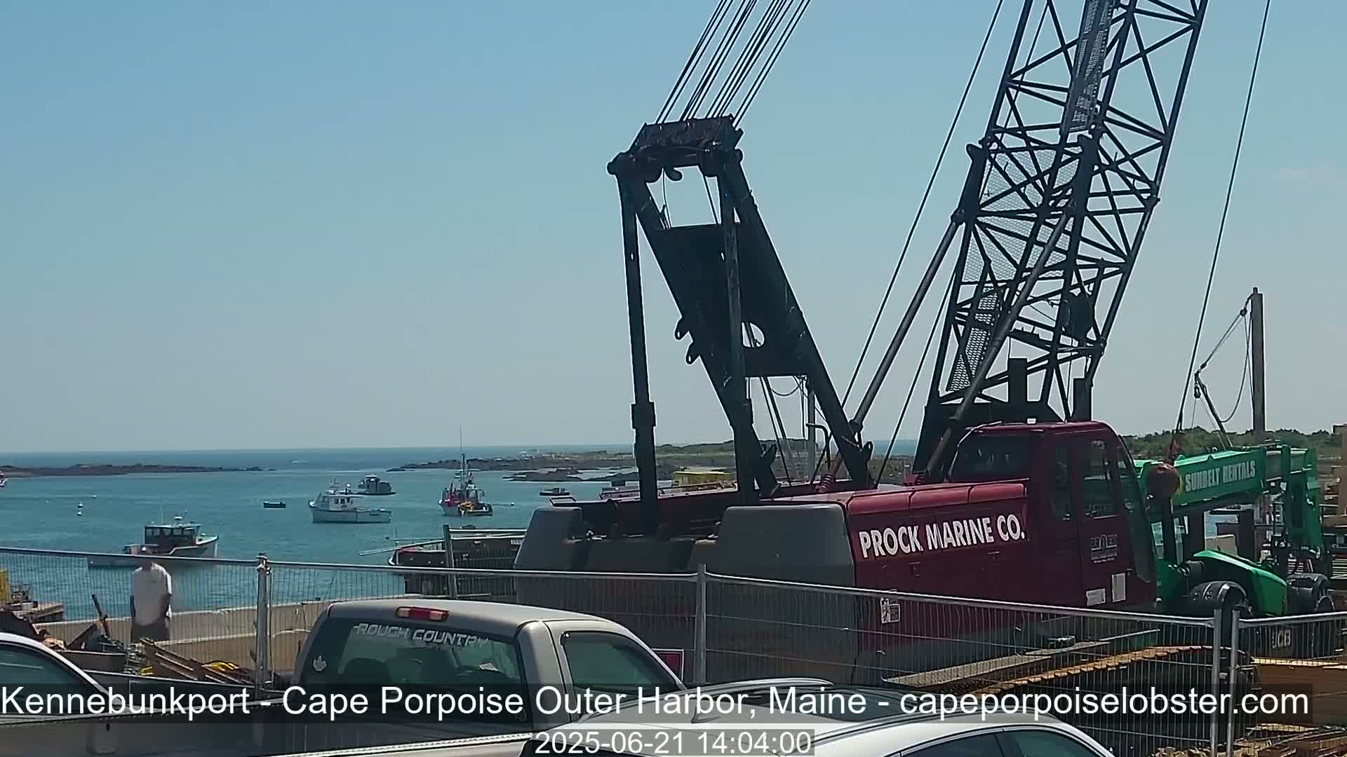 A large red crane is positioned near a harbor on a sunny day, with several fishing boats visible on the calm water in the background.