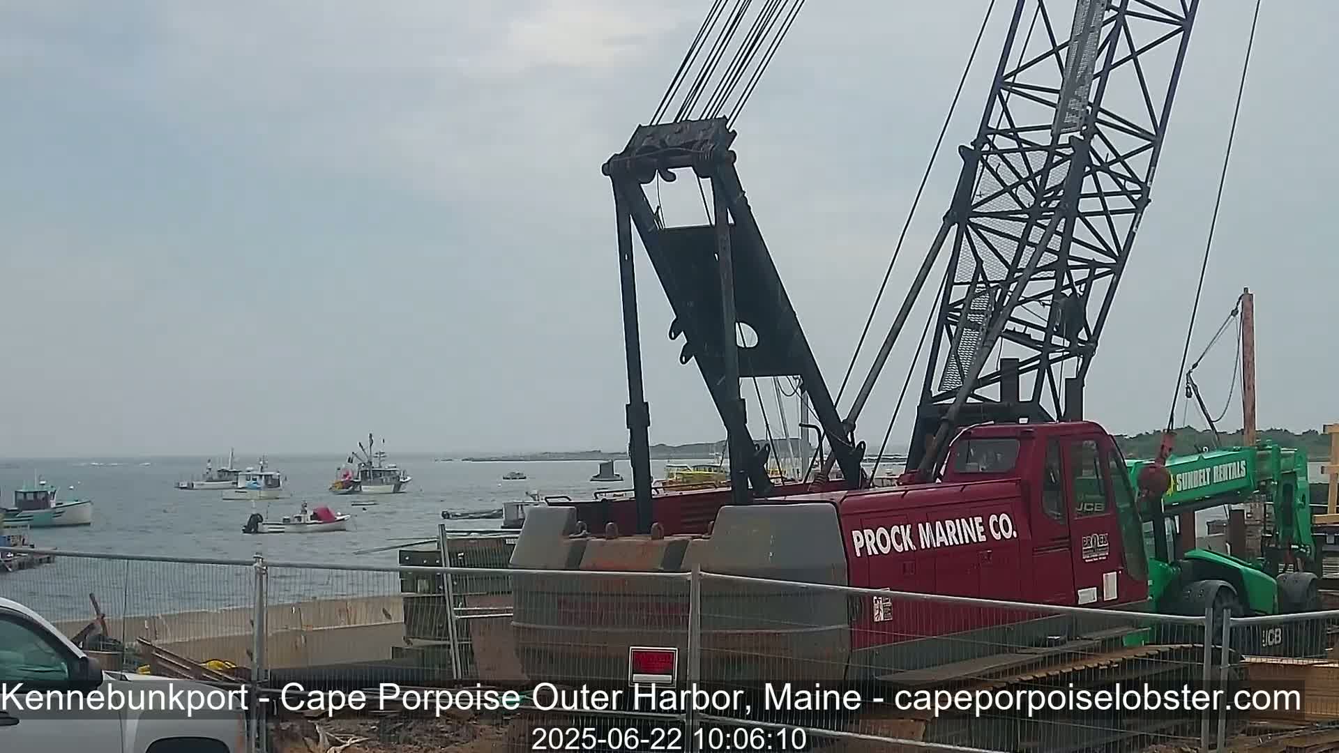 A red Prock Marine Co. crane is positioned near a harbor with several small boats on an overcast day.