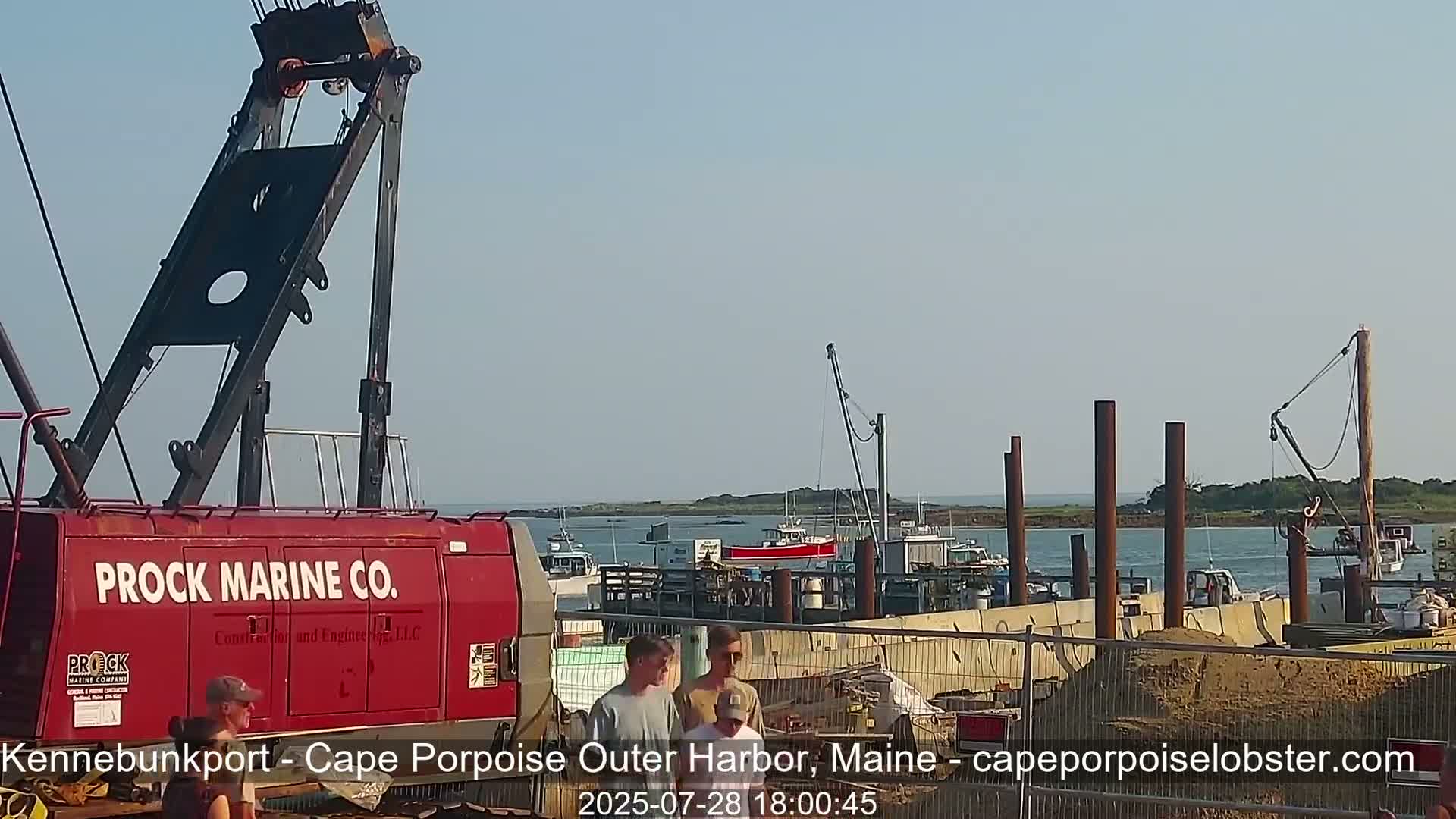 A red Prock Marine Co. crane is parked near a harbor where construction is underway under a clear sky.