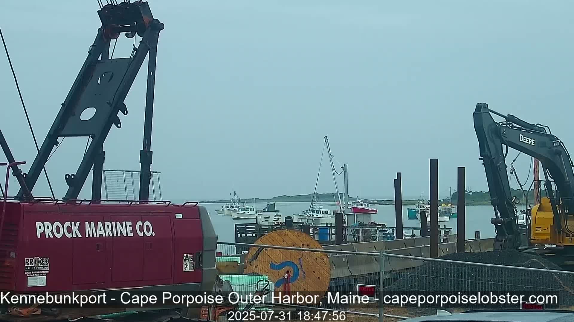 A red crane, a gray excavator, and several small boats are situated at a harbor under an overcast sky.
