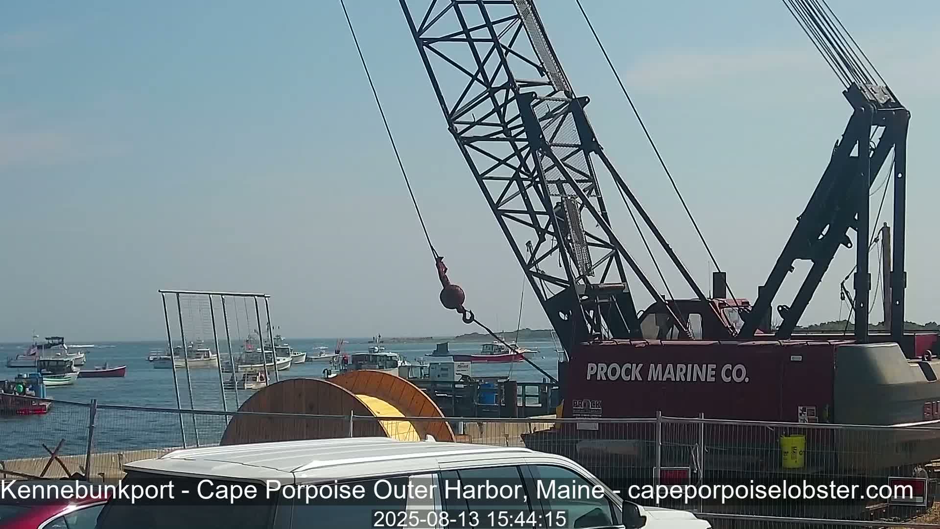 A large crane stands near a harbor on a sunny day, with numerous small boats in the water.