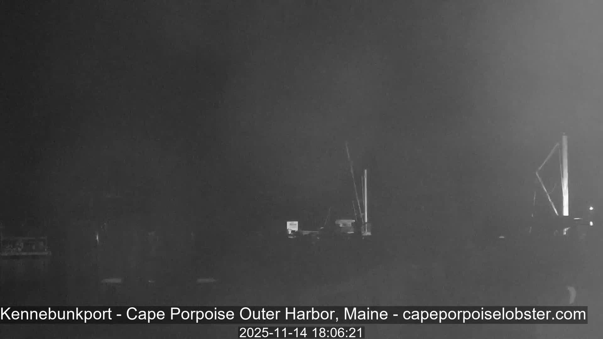 A very dark outdoor scene, likely a harbor at night, dimly shows faint vertical structures such as boat masts or dock pilings, with extremely low visibility suggesting a foggy or heavily overcast atmosphere.