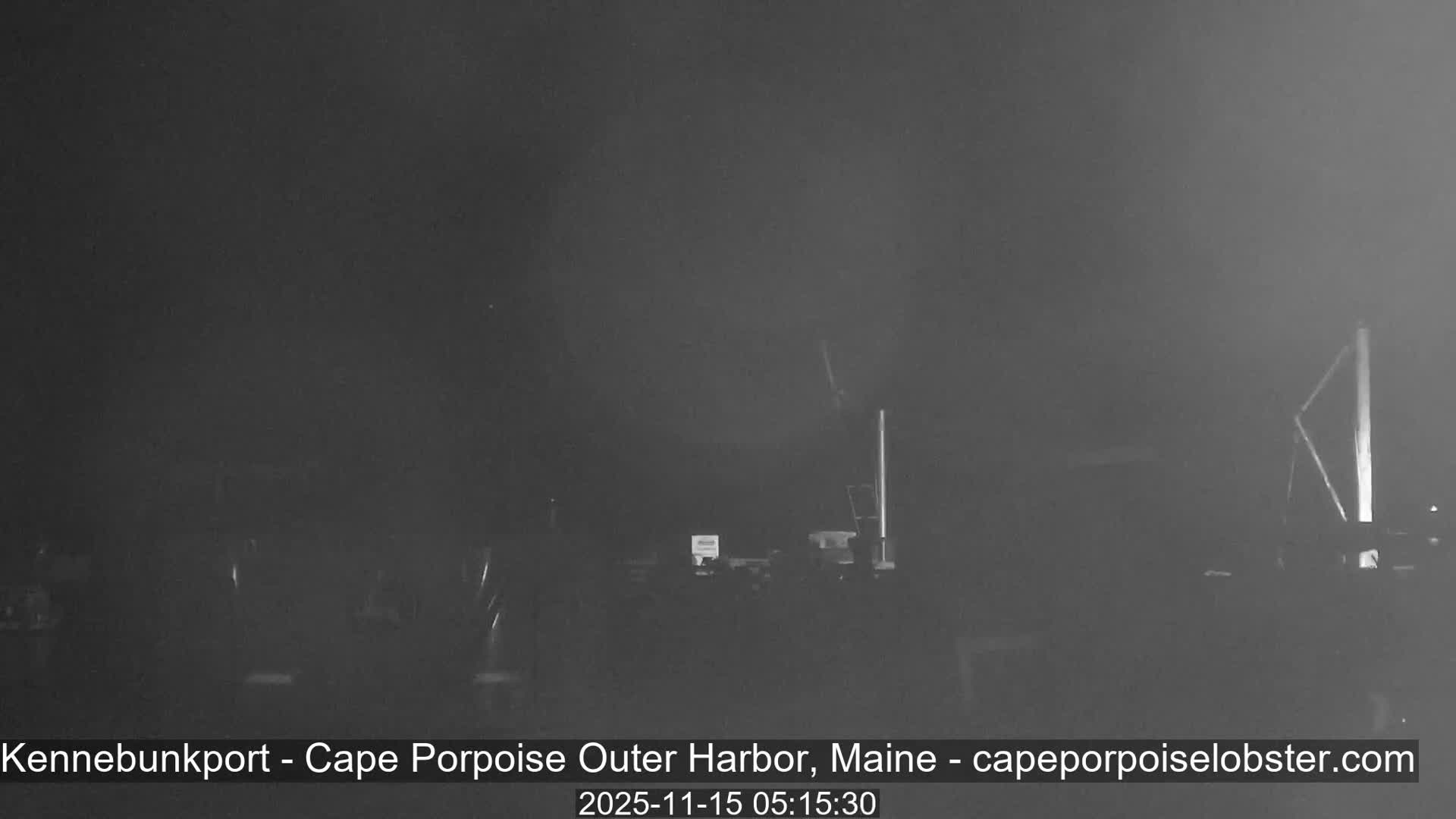 The image shows a dark and heavily fog-obscured outdoor scene, likely a harbor, with only faint outlines of upright poles and some structures discernible through the very low visibility conditions.