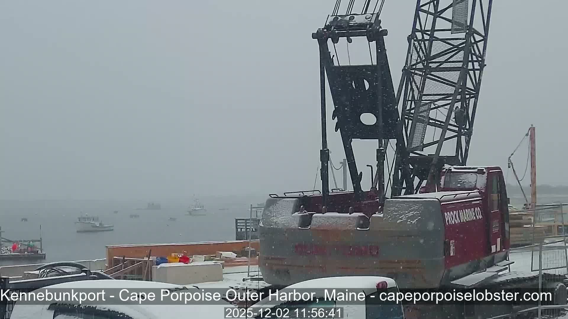 Heavy snow falls over a harbor where a large red and gray crane stands partially covered in snow on the right, with numerous fishing boats scattered across the gray water on the left.