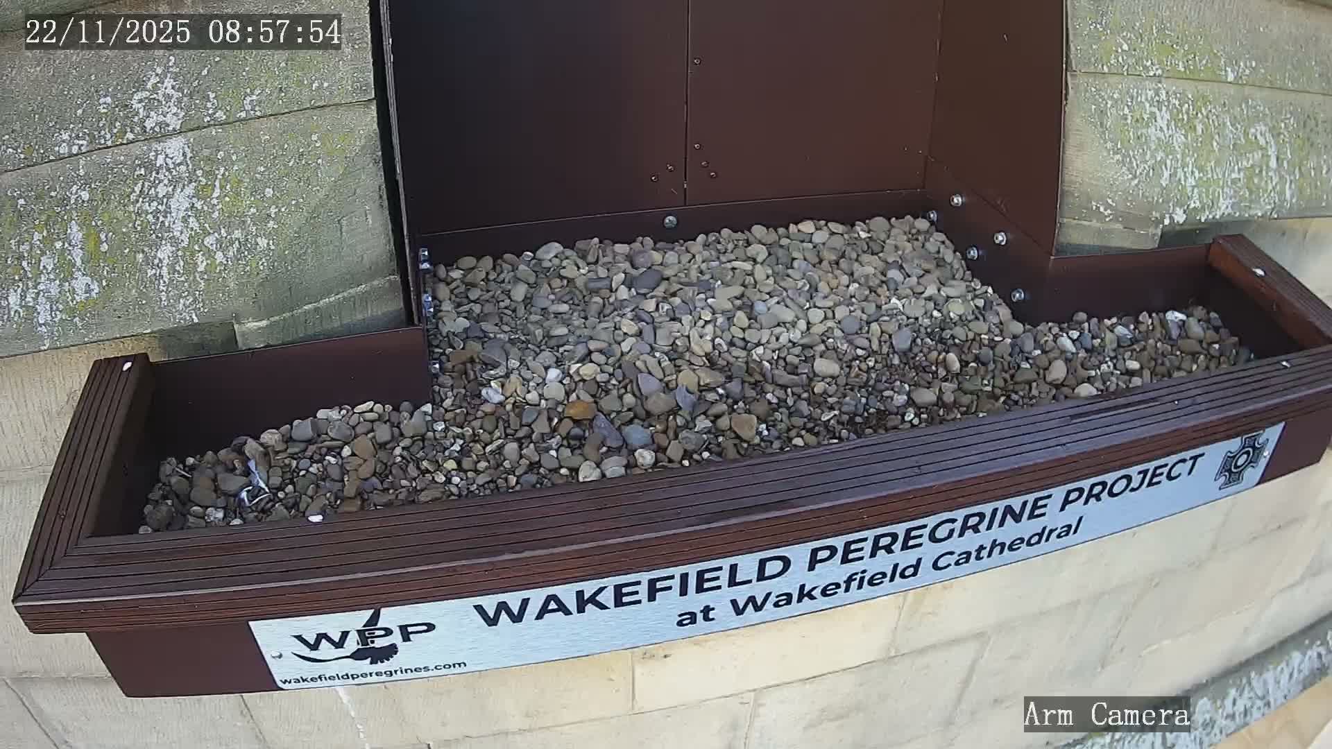 A dark brown, two-tiered structure filled with various-colored pebbles is attached to a textured, light stone wall under bright, clear daylight.