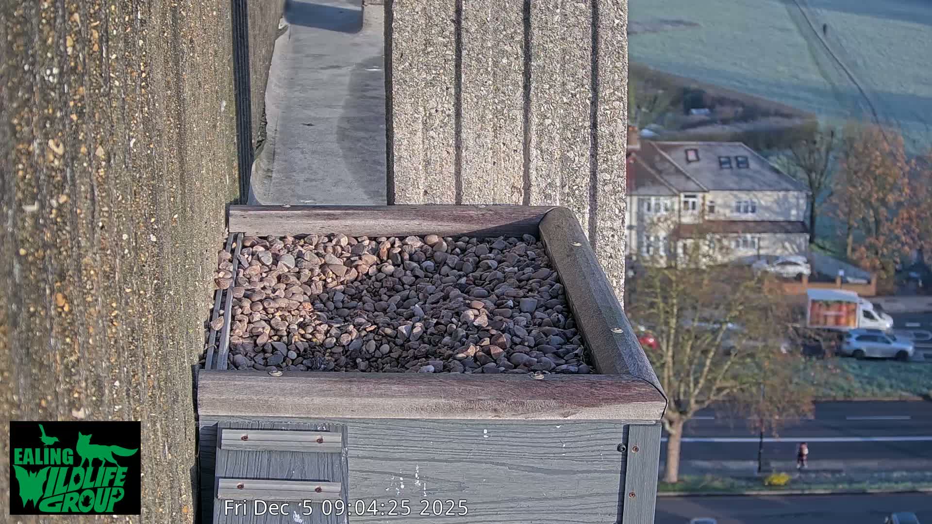 A wooden box filled with pebbles sits on a building ledge, overlooking a frosty suburban landscape with houses, bare trees, and a road on a clear, cold morning.