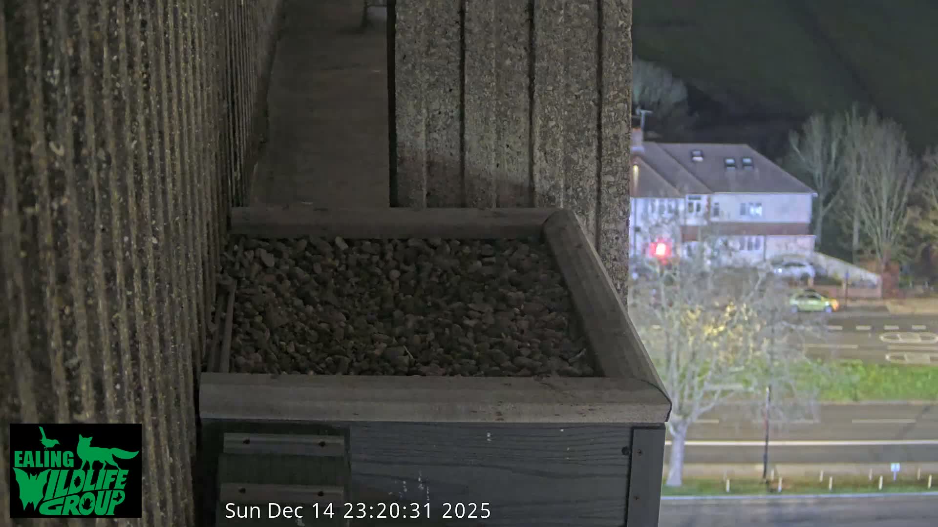 A wooden box filled with pebbles sits on a building ledge, overlooking a frosty suburban landscape with houses, bare trees, and a road on a clear, cold morning.
