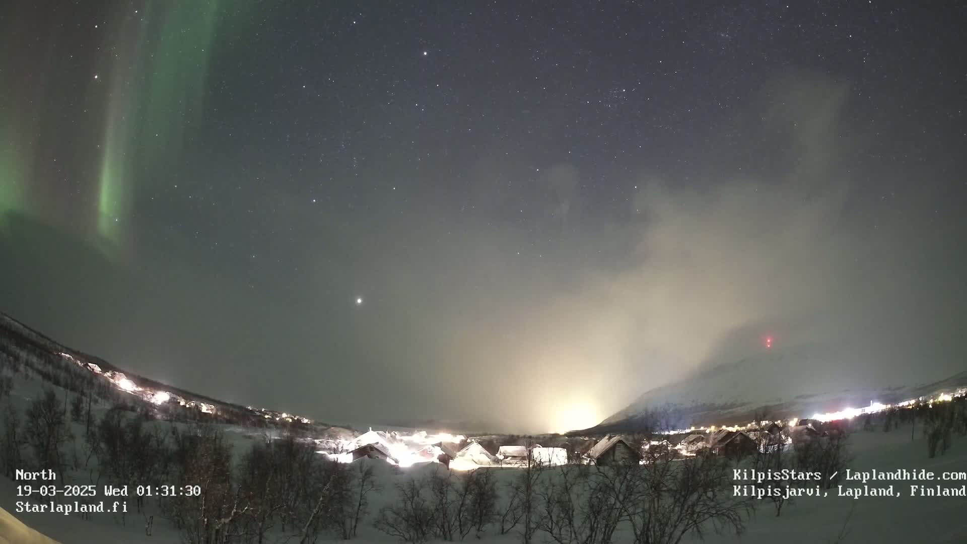 A snowy village is illuminated at night under a starry sky displaying a vibrant aurora borealis and a hazy, light-colored glow near a mountain.