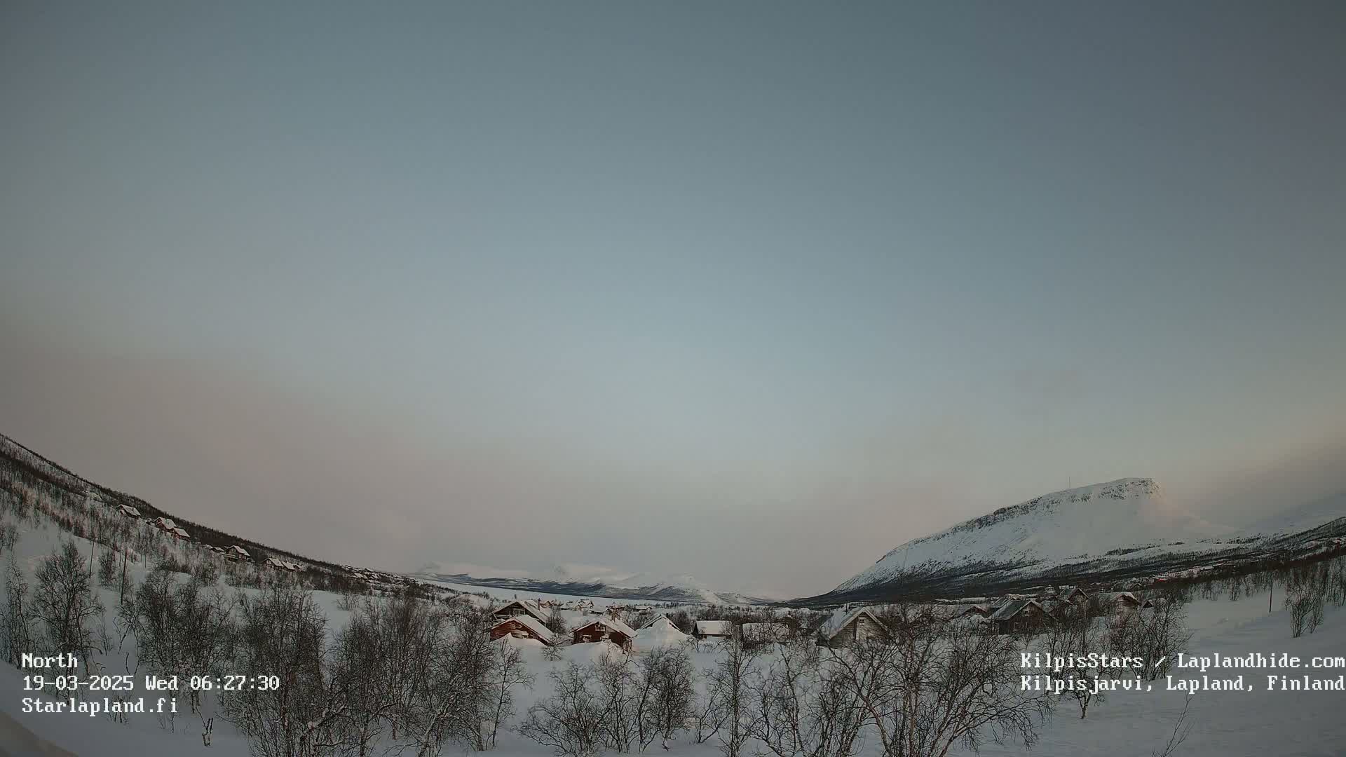 A snow-covered landscape with small houses nestled among leafless trees, mountains in the background under a clear, pale sky.