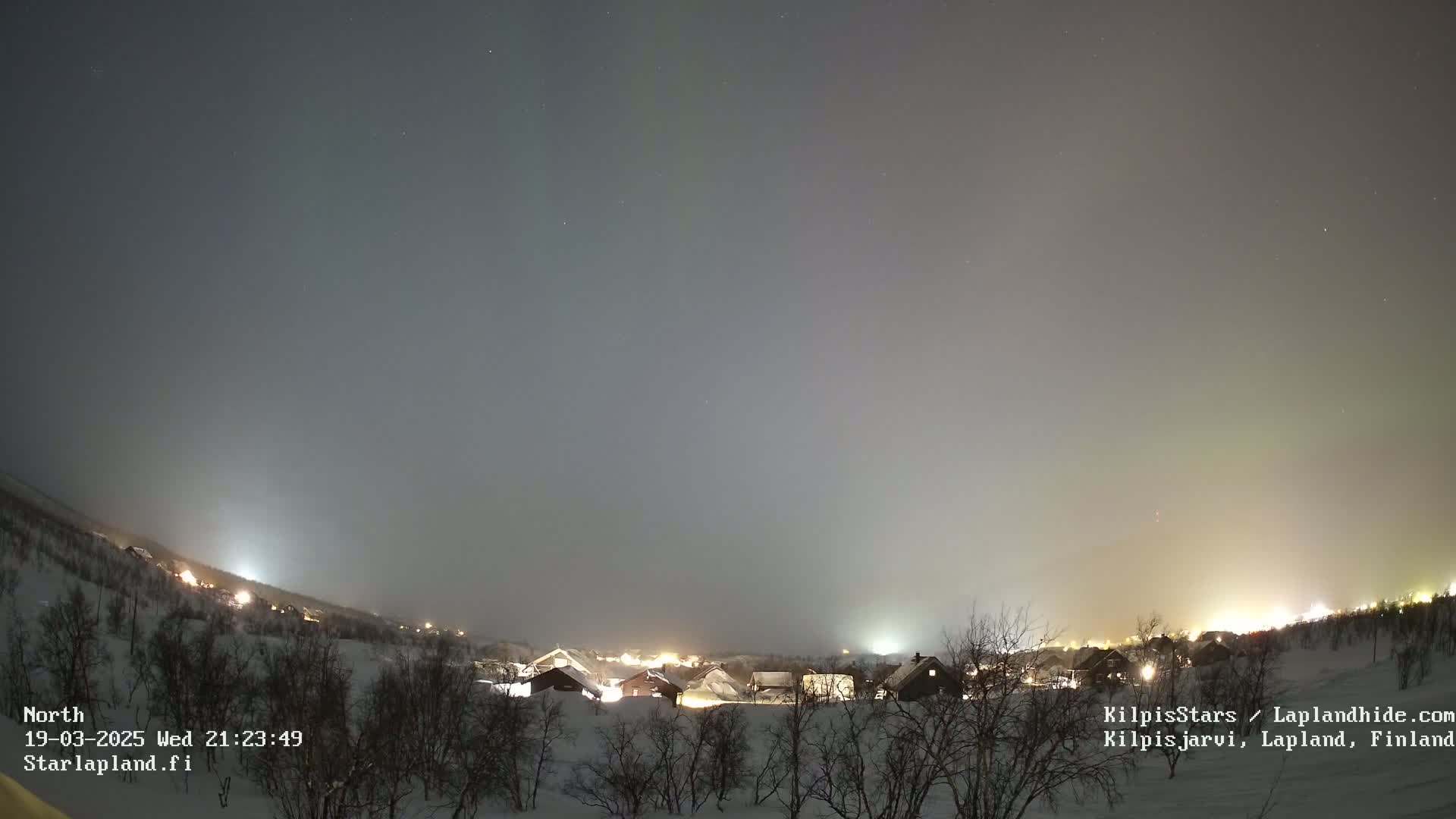 A faint aurora borealis illuminates a night sky over a snow-covered village with sparsely wooded hills in the distance.