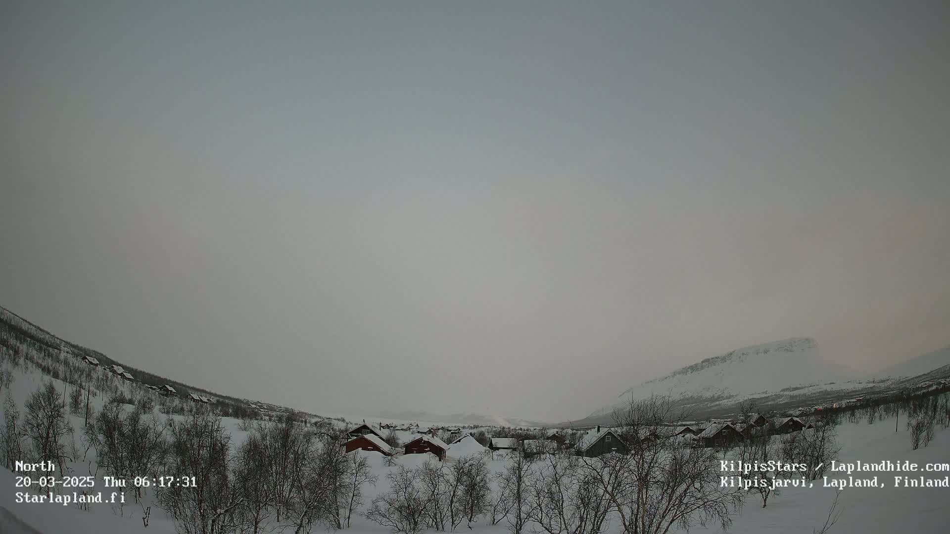 A snow-covered landscape with a village of small houses nestled in a valley between snow-dusted mountains under an overcast sky.