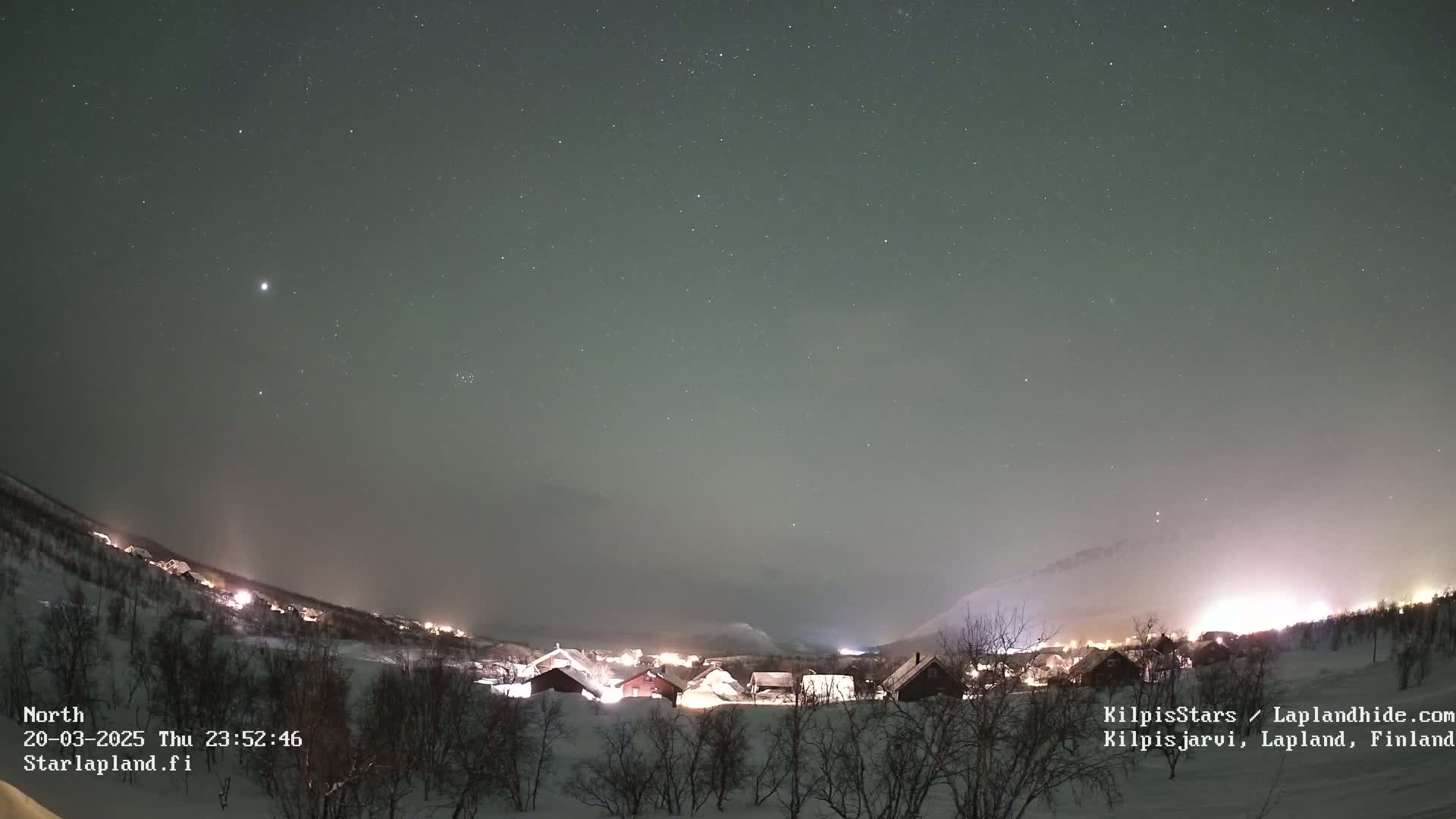 A snow-covered village is illuminated at night under a starry sky with a faint aurora borealis.