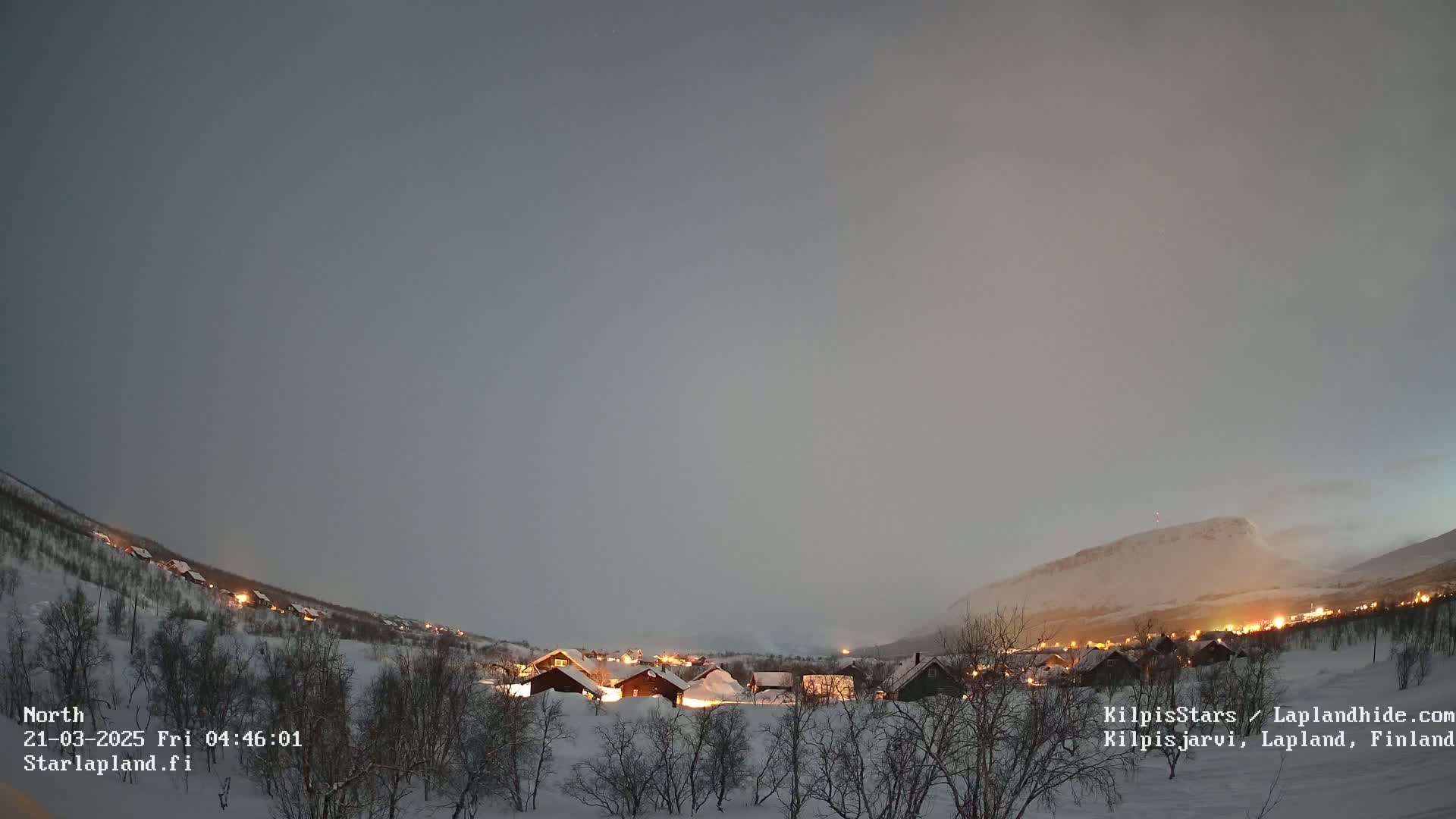 A snow-covered village with illuminated houses sits at the base of a snow-capped mountain under a twilight sky.