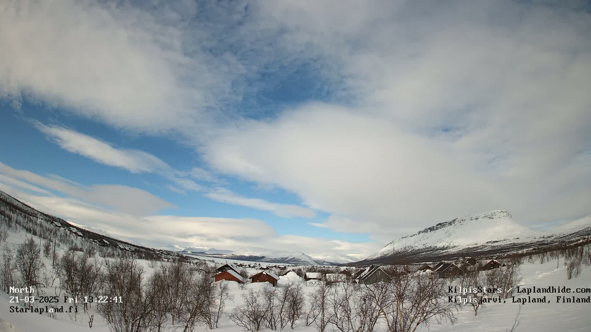 A snow-covered village nestled in a valley between snow-capped mountains under a partly cloudy sky.