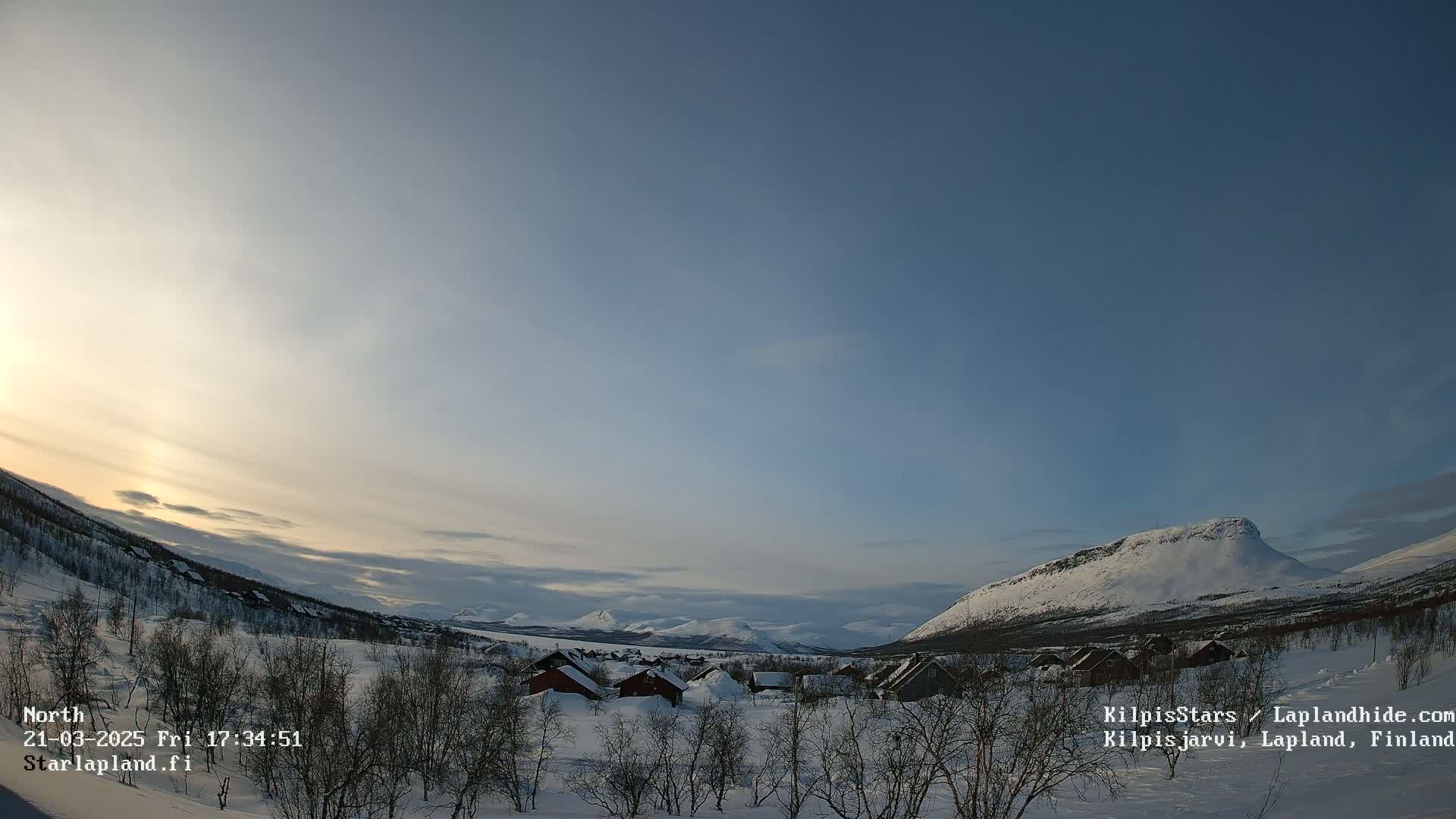 A snow-covered landscape with a village of small houses nestled in a valley, under a mostly clear, twilight sky.