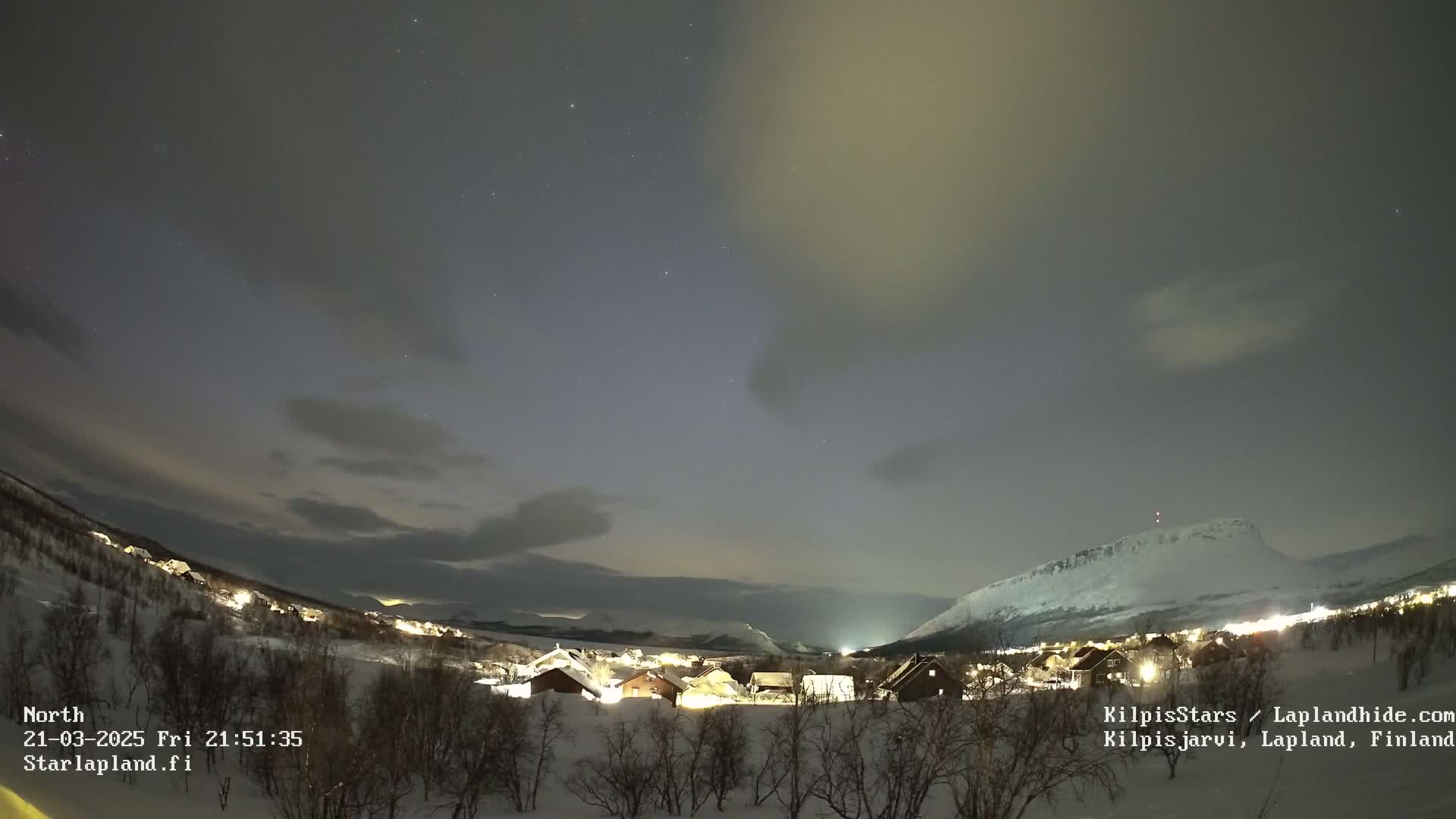 A nighttime, snowy landscape shows a small village nestled below a snow-covered mountain, with a faint aurora borealis visible in the night sky.