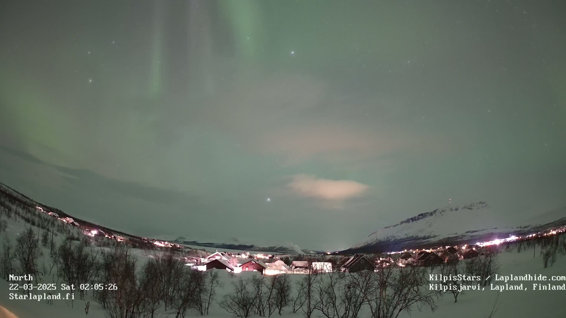 A snow-covered village is illuminated at night under a pale green aurora borealis in a mostly clear sky.