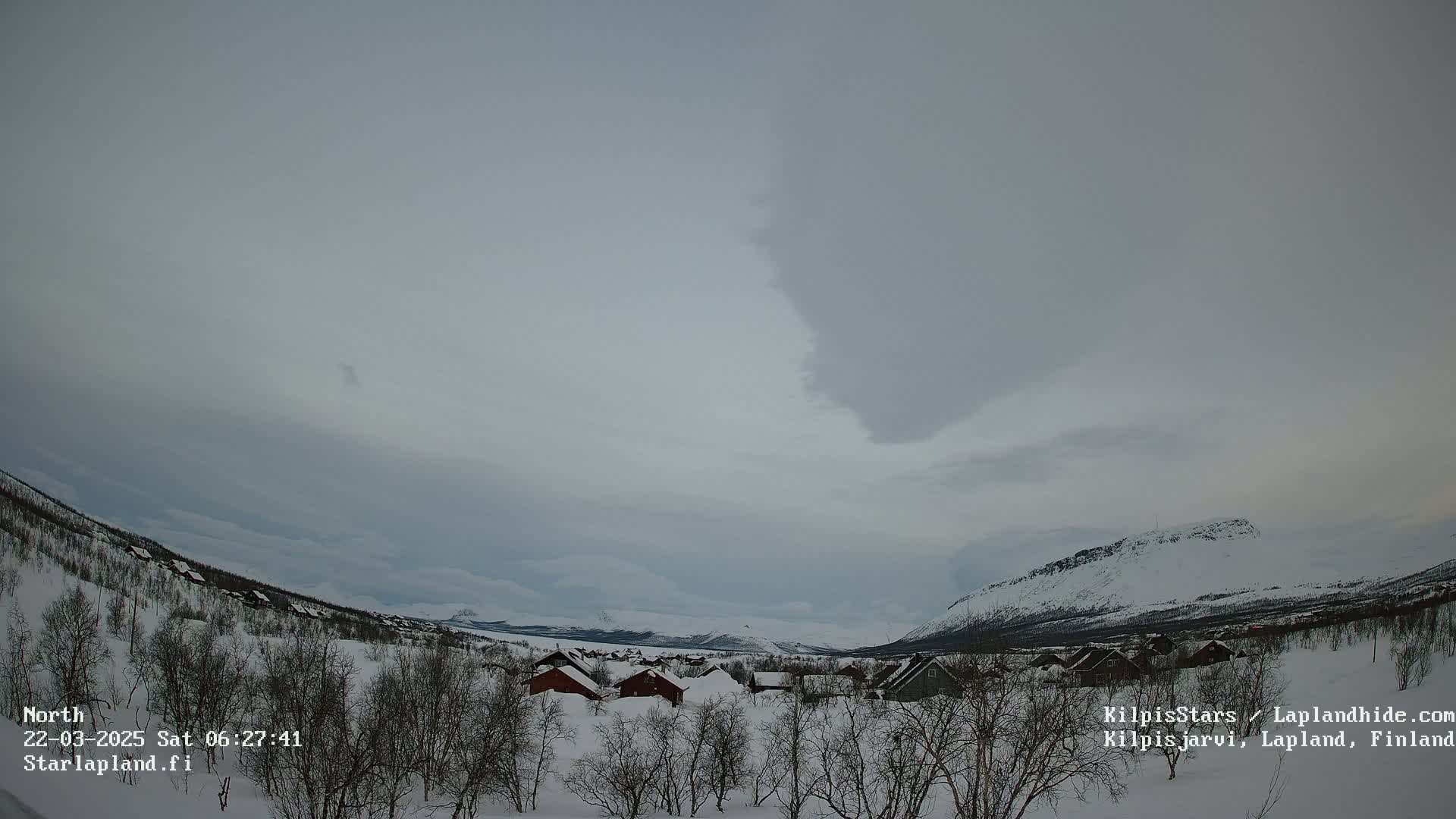 A snow-covered landscape with a small village nestled amongst leafless trees, under a mostly cloudy, overcast sky.