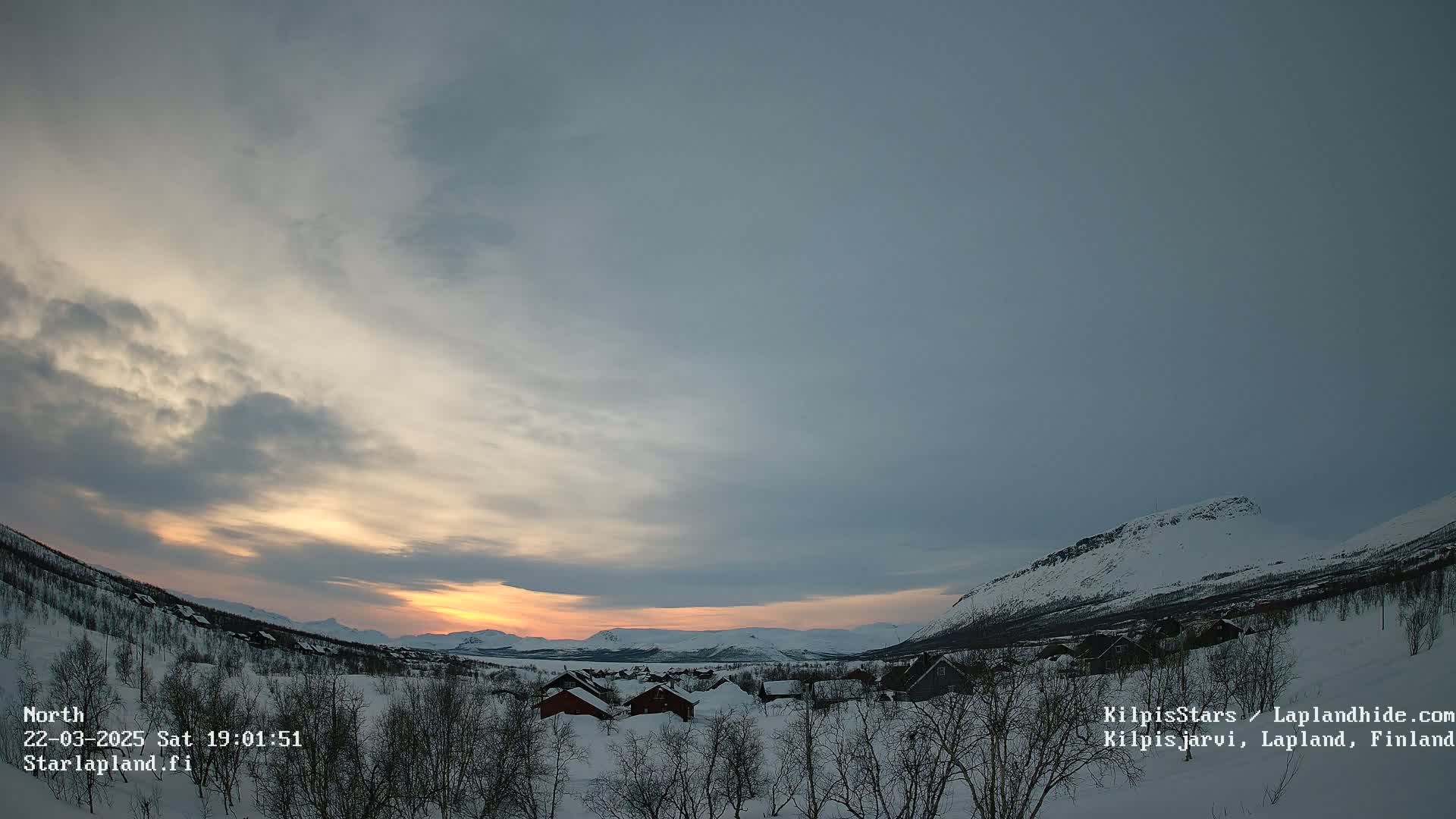 A snow-covered village nestled in a valley between mountains, bathed in the soft light of a sunset under a mostly cloudy sky.