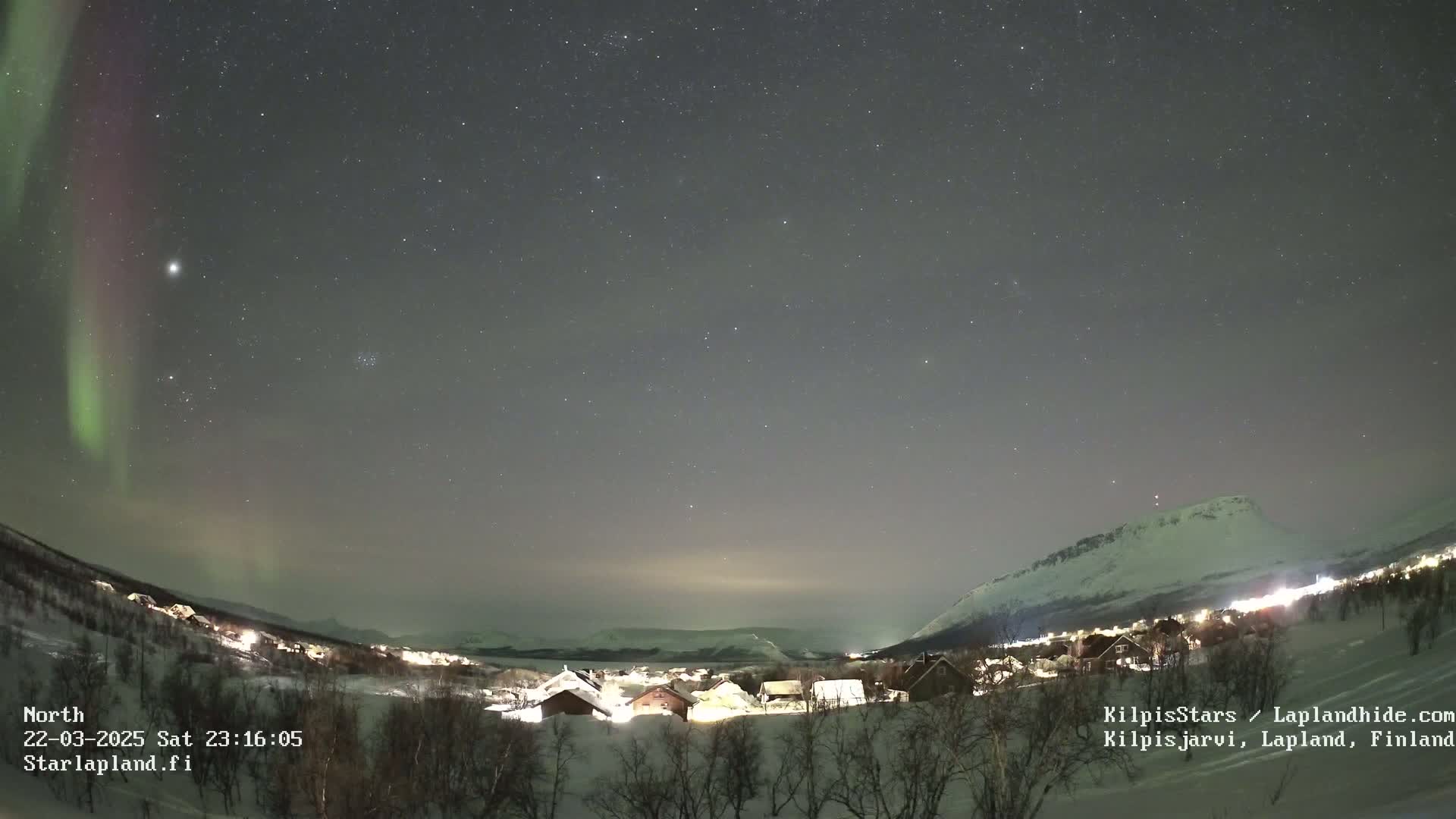 A snow-covered village is illuminated at night under a dark sky filled with stars and a vibrant aurora borealis.