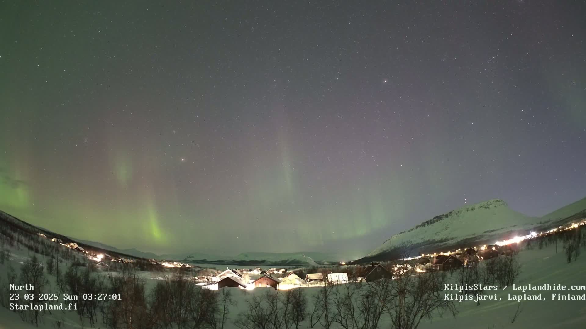 A nighttime view of a snowy village nestled in a valley beneath a vibrant aurora borealis display against a starry sky.