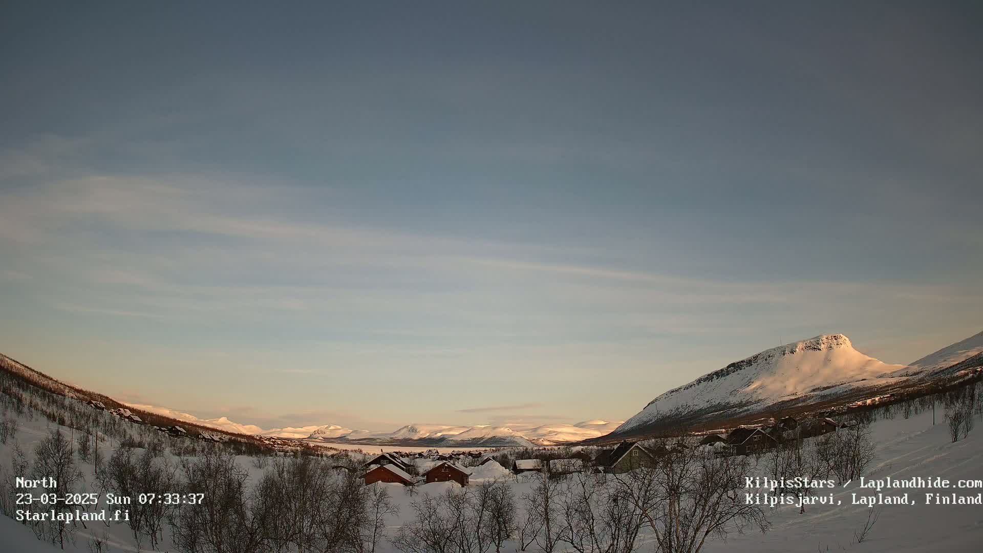 A snow-covered landscape with a small village nestled among the foothills of a mountain range under a pale, clear sky.
