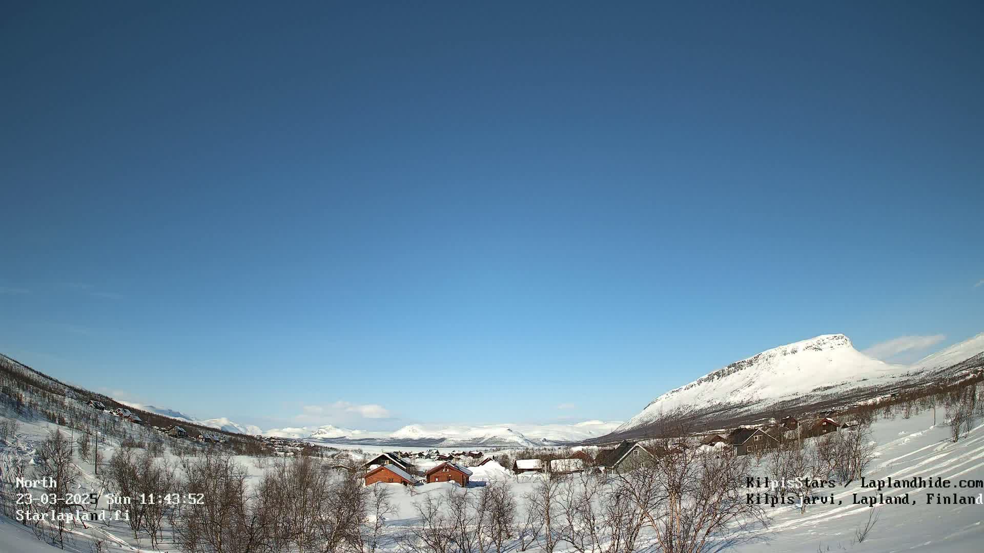 A snow-covered village nestled in a valley between snow-capped mountains under a clear blue sky.